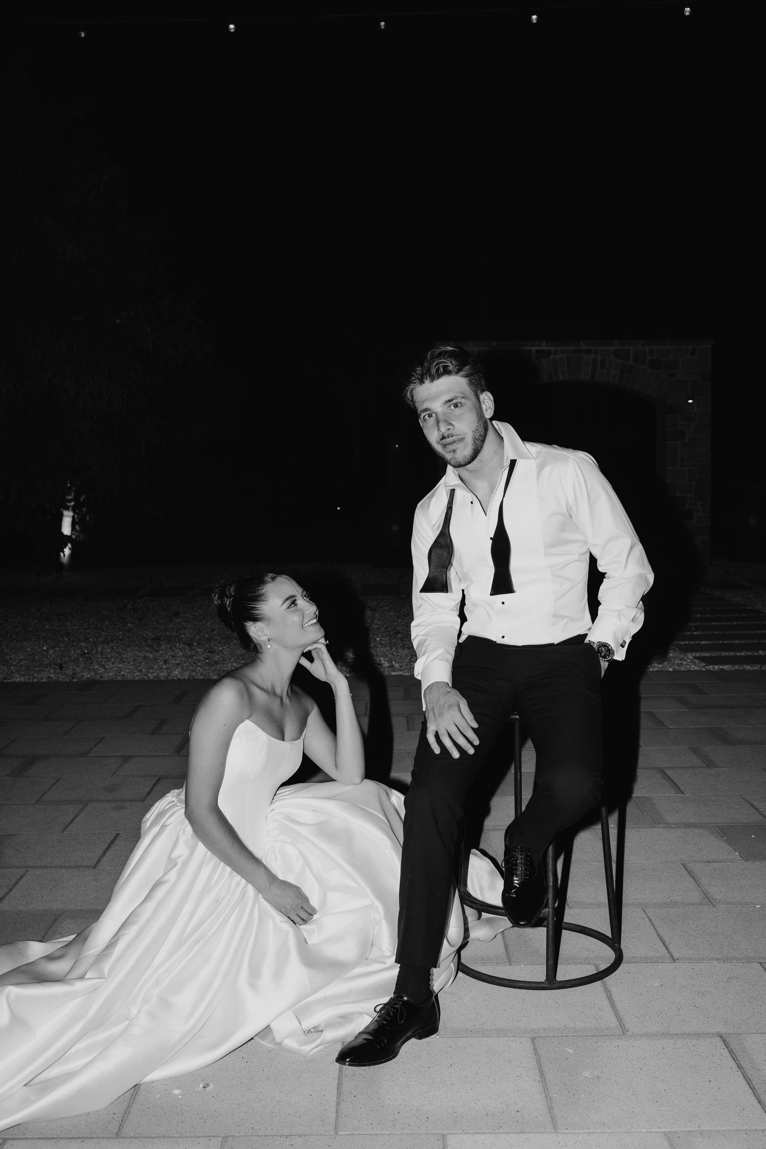 Black and white photo of a bride sitting on the floor and groom seated on a stool at an outdoor evening wedding.