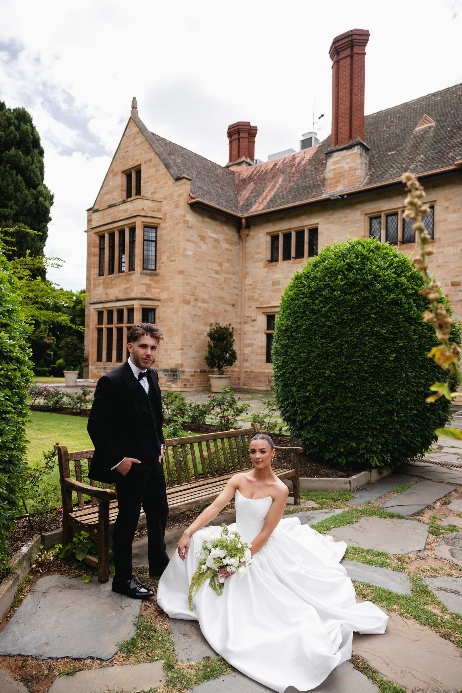 Bride in white gown sits on stone steps holding bouquet while groom in black tux stands beside her at elegant manor.