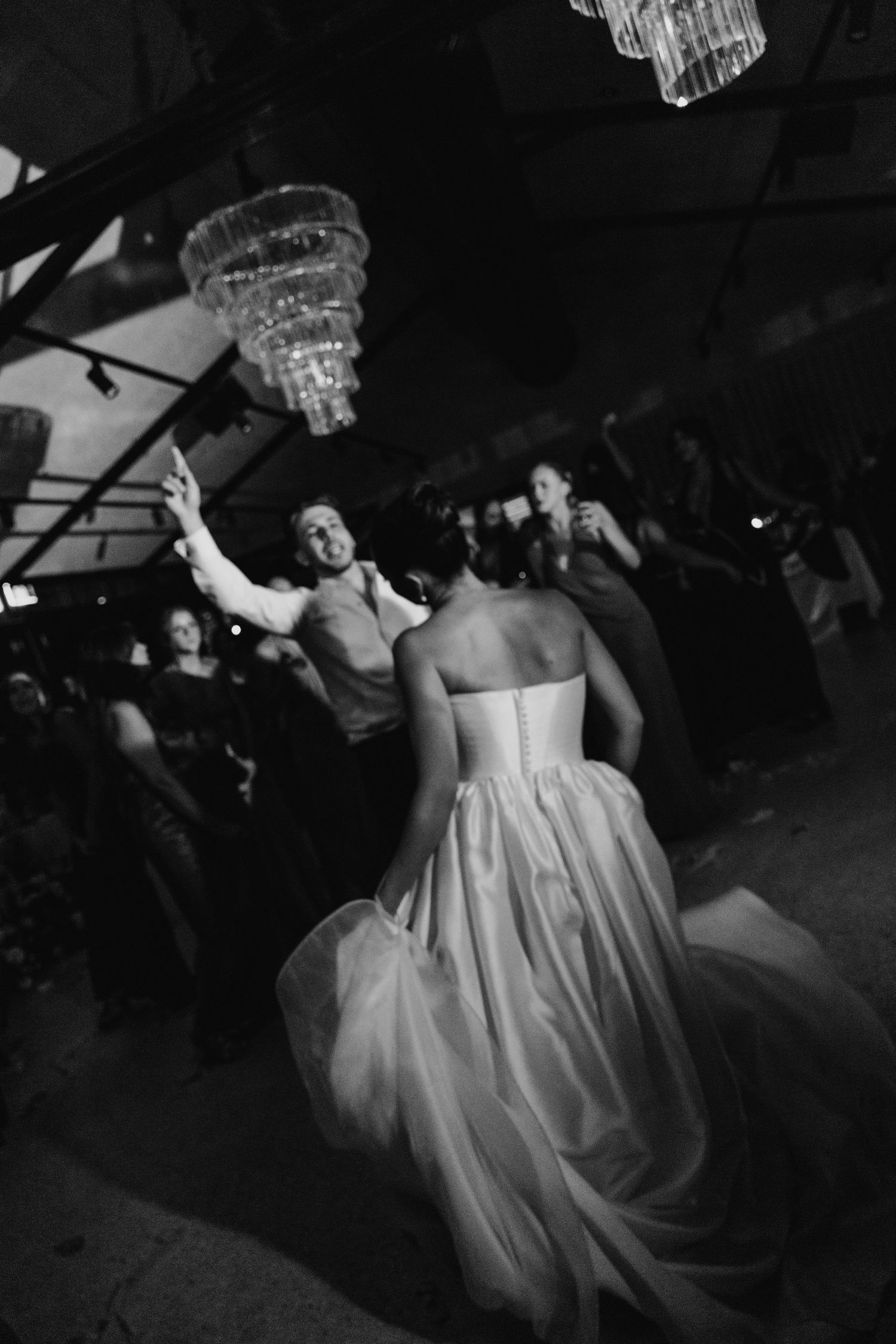 Black and white photo of bride and groom sharing first dance under chandelier at elegant wedding reception.