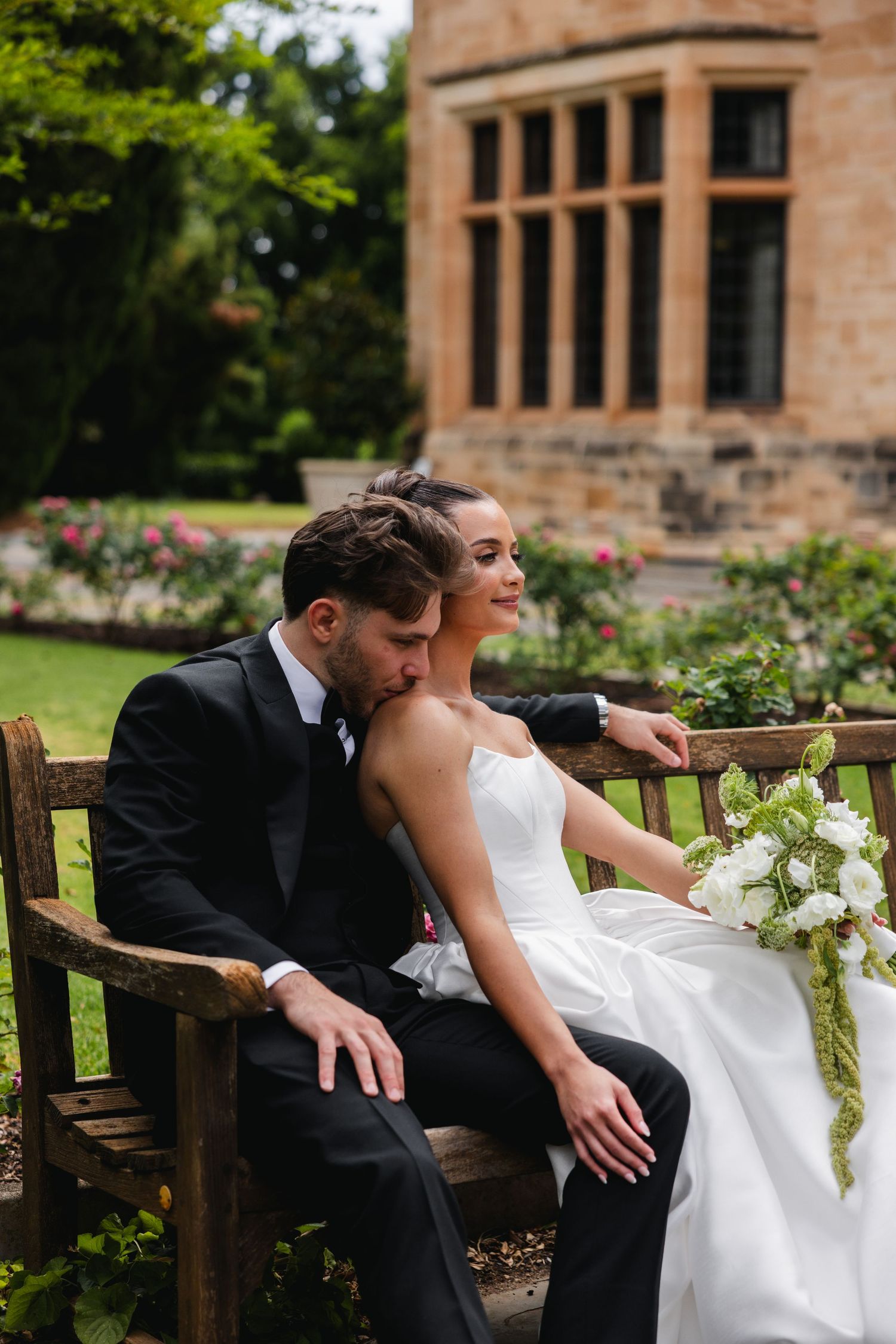 Bride and groom sitting on a wooden bench in a garden, groom kissing bride's cheek, bride holding white bouquet.