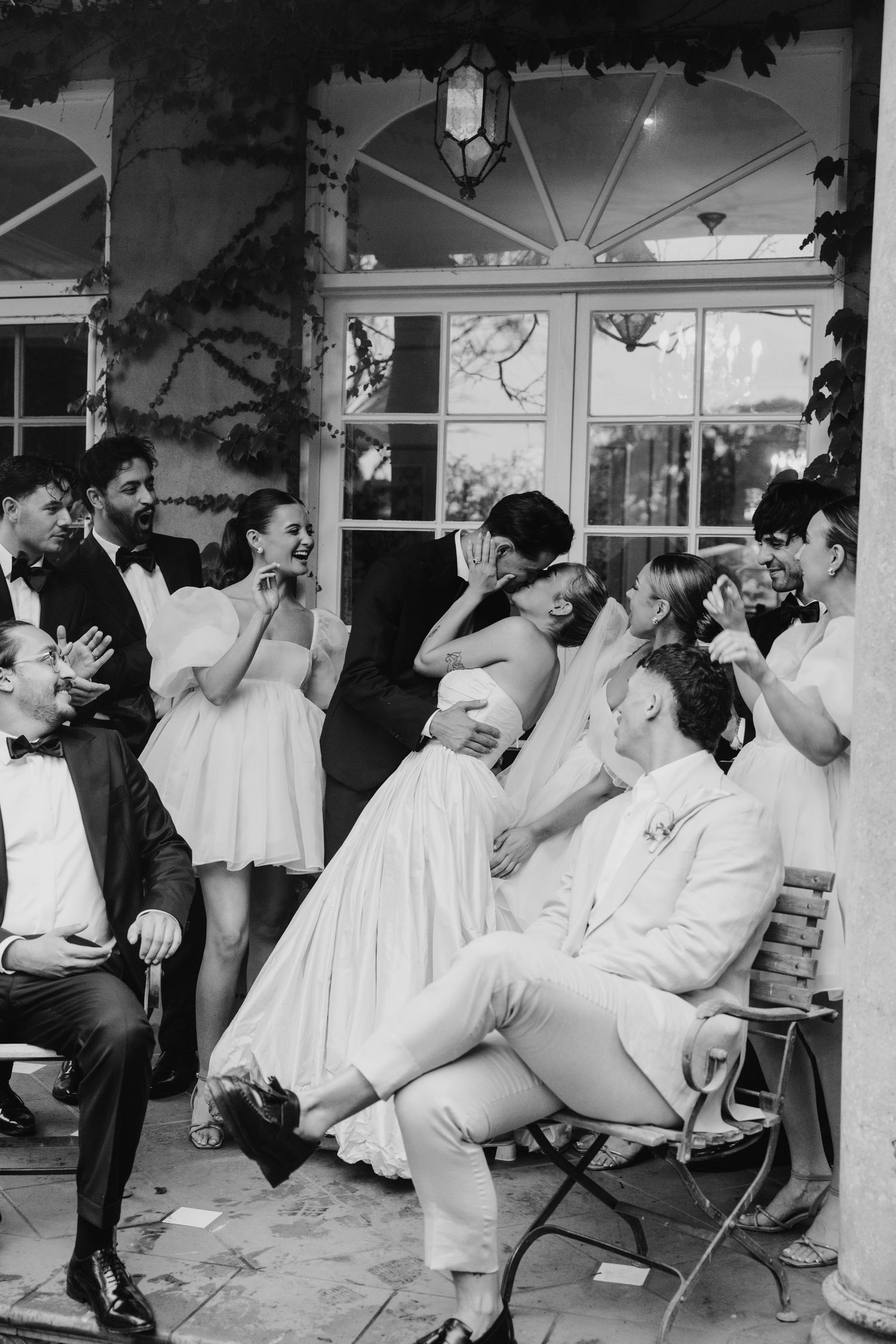 Joyful wedding party candid moment with bride and groom kissing on chair in black and white.