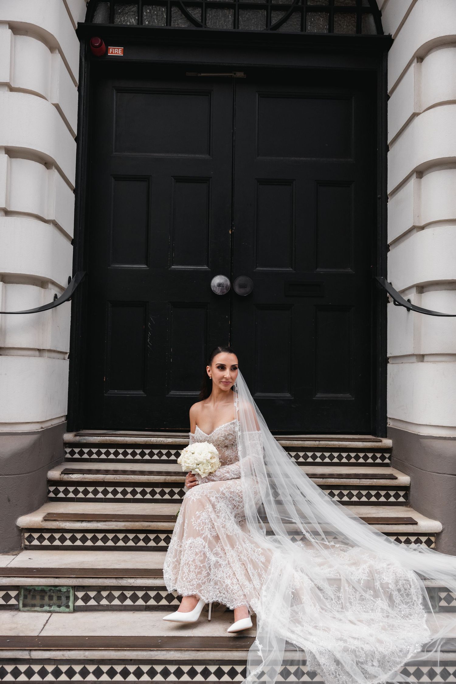 Elegant bride in lace wedding dress and veil sitting on tiled steps in front of a large black door.