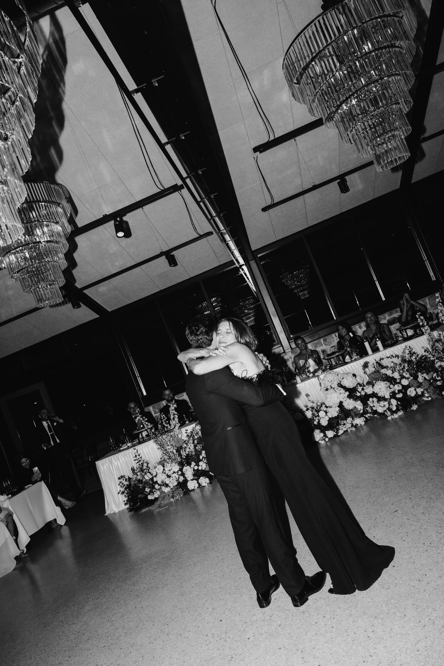 Black and white photo of two people sharing an embrace on a dance floor at an elegant wedding reception with chandeliers.