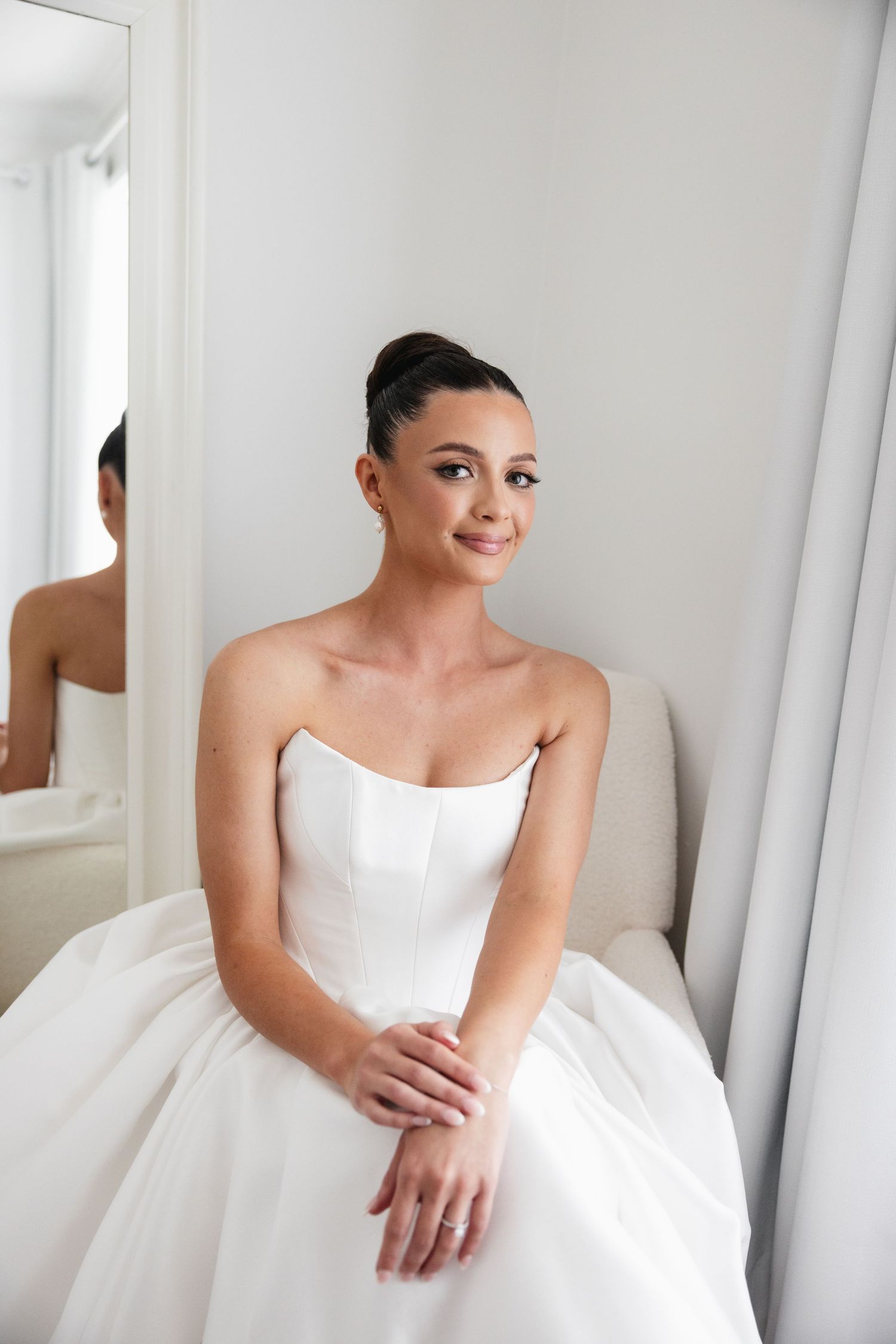 Elegant bride in strapless white wedding gown sitting gracefully beside a mirror in a bright bridal suite.
