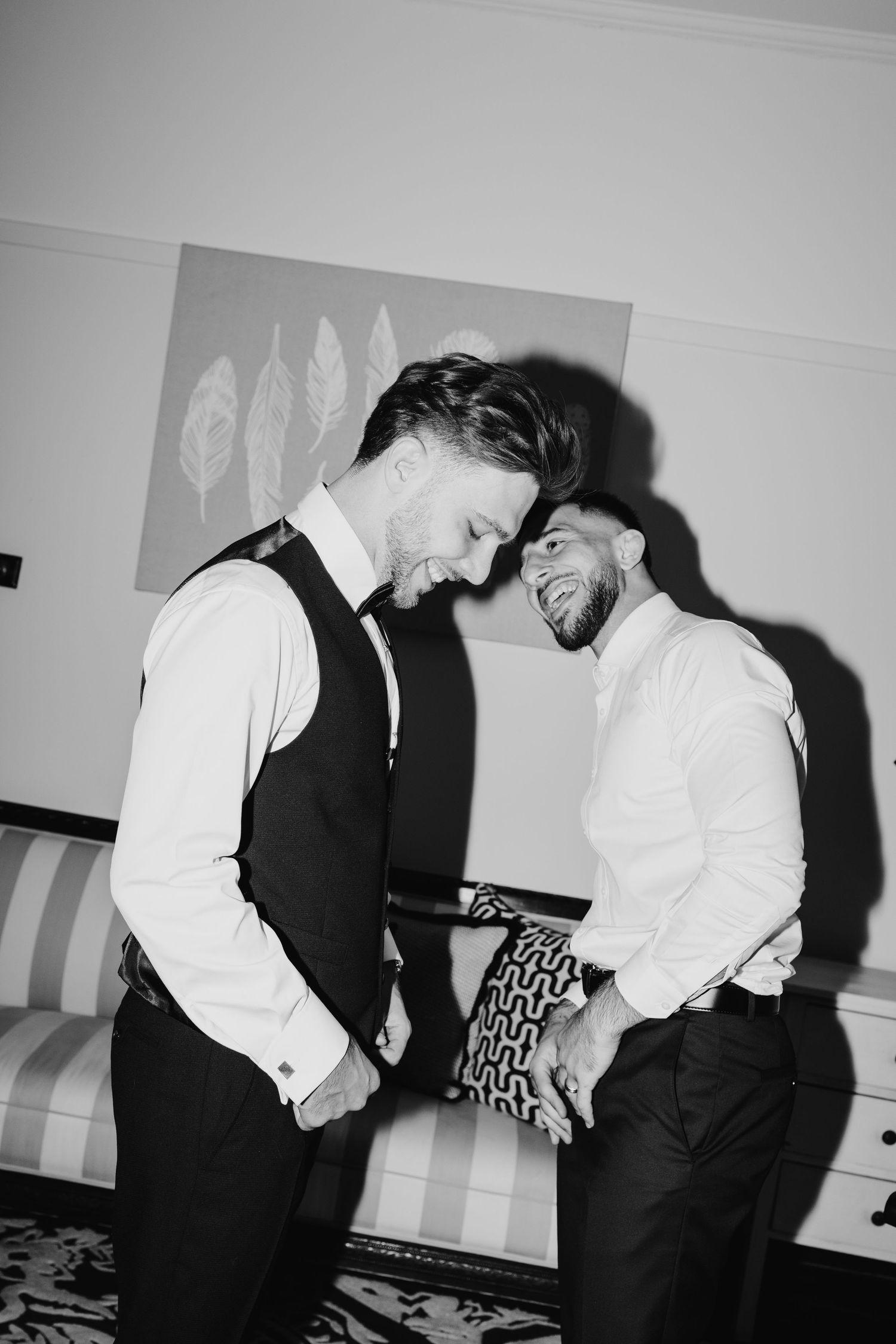 Black and white photo of two men in formal attire smiling and holding hands together indoors.
