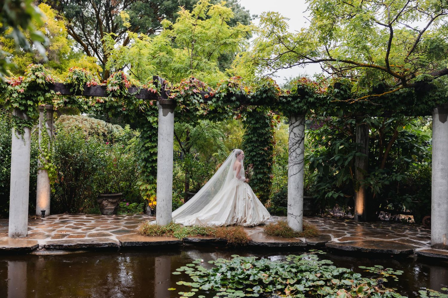 Bride in flowing white gown and veil stands beside a lily pond under a lush garden pergola with white columns.