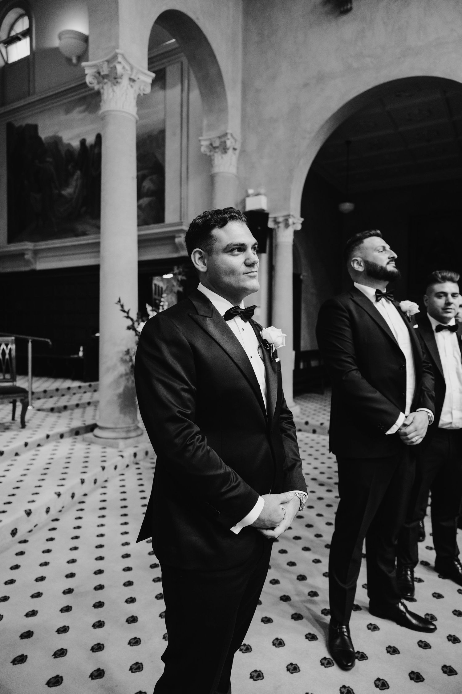 Groom in black tuxedo waiting at altar with groomsmen in elegant venue.