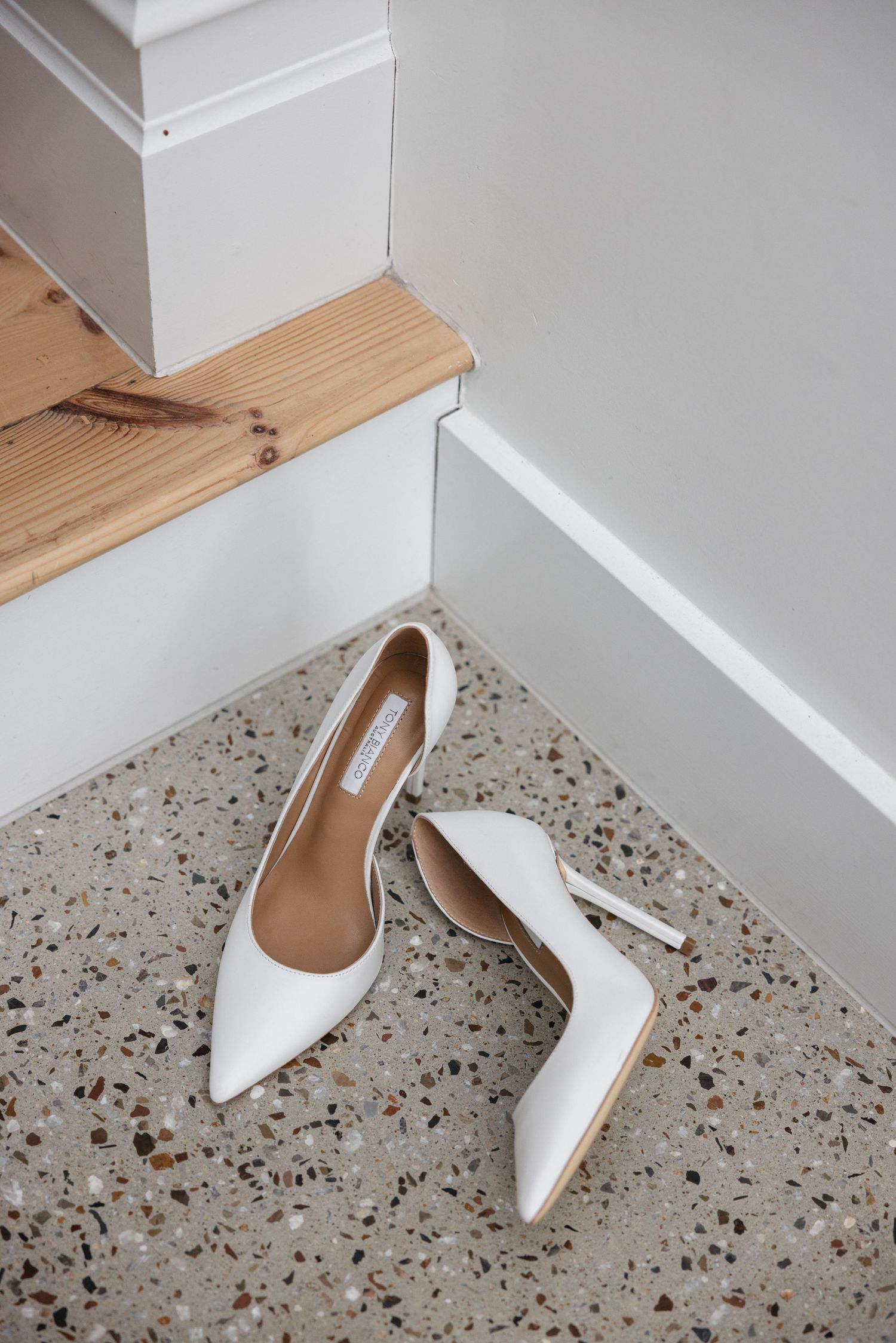 White stiletto heels placed on terrazzo floor beside wooden stairs, perfect bridal shoes for a wedding day.