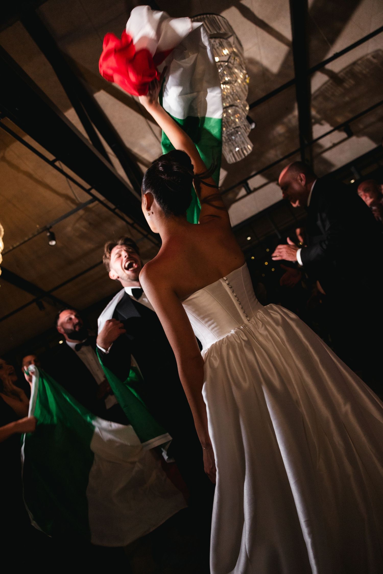 Bride in white strapless gown waves Italian flag at wedding reception celebration with guests in formal attire.