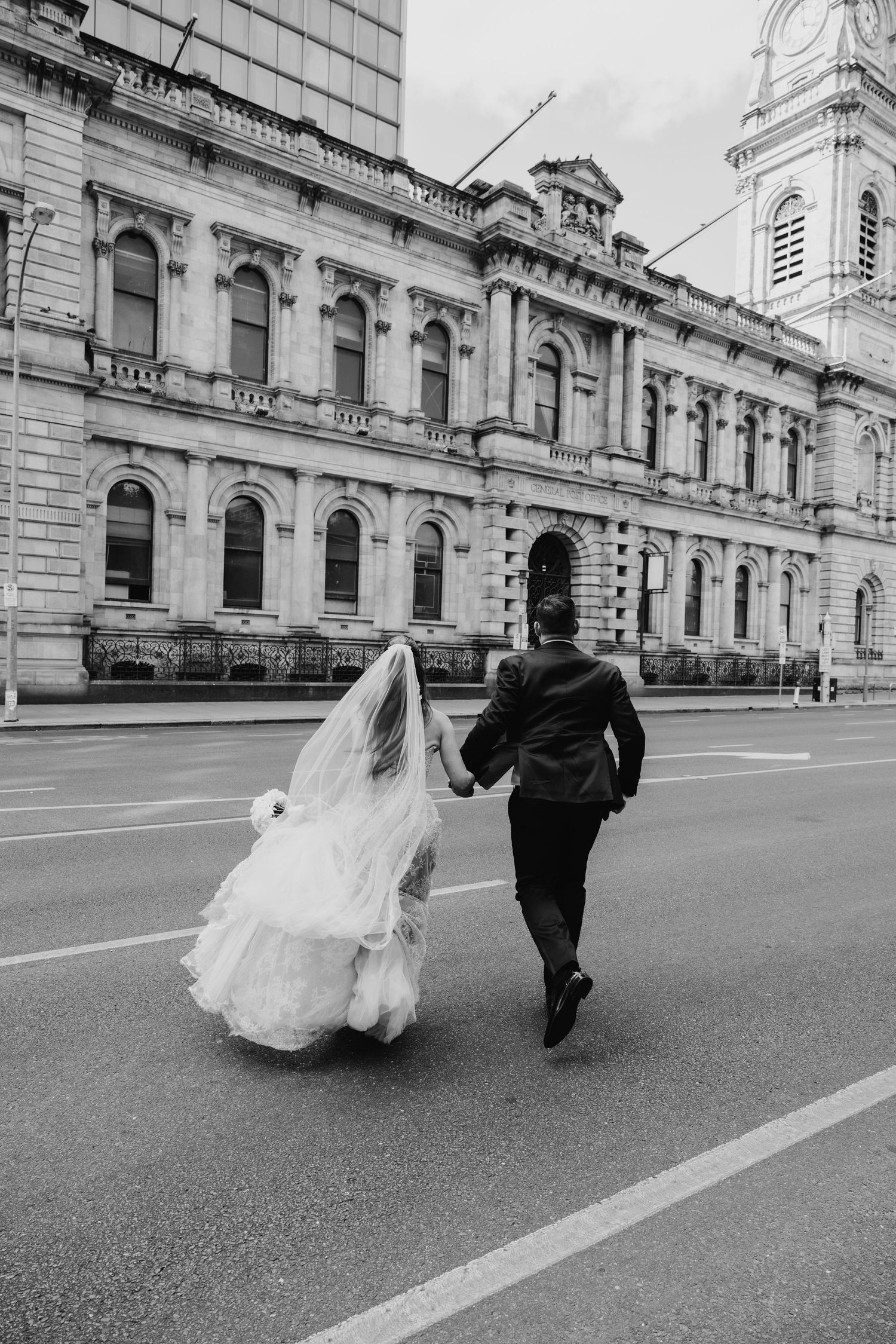 Bride and groom running hand in hand across street near historic building.