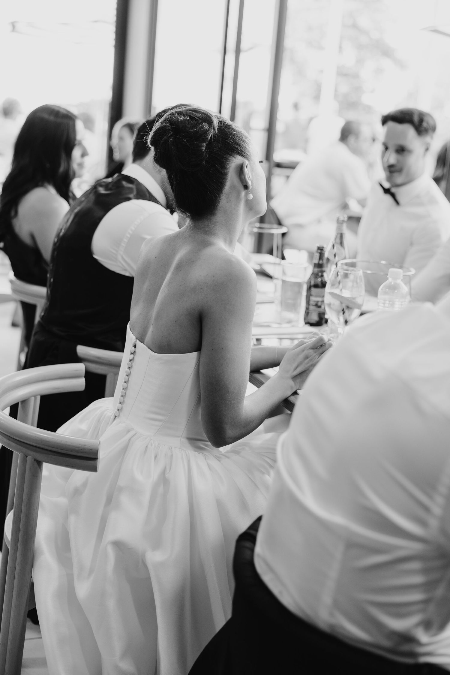 Black and white photo of a bride in a strapless gown seated at a wedding reception table with guests.