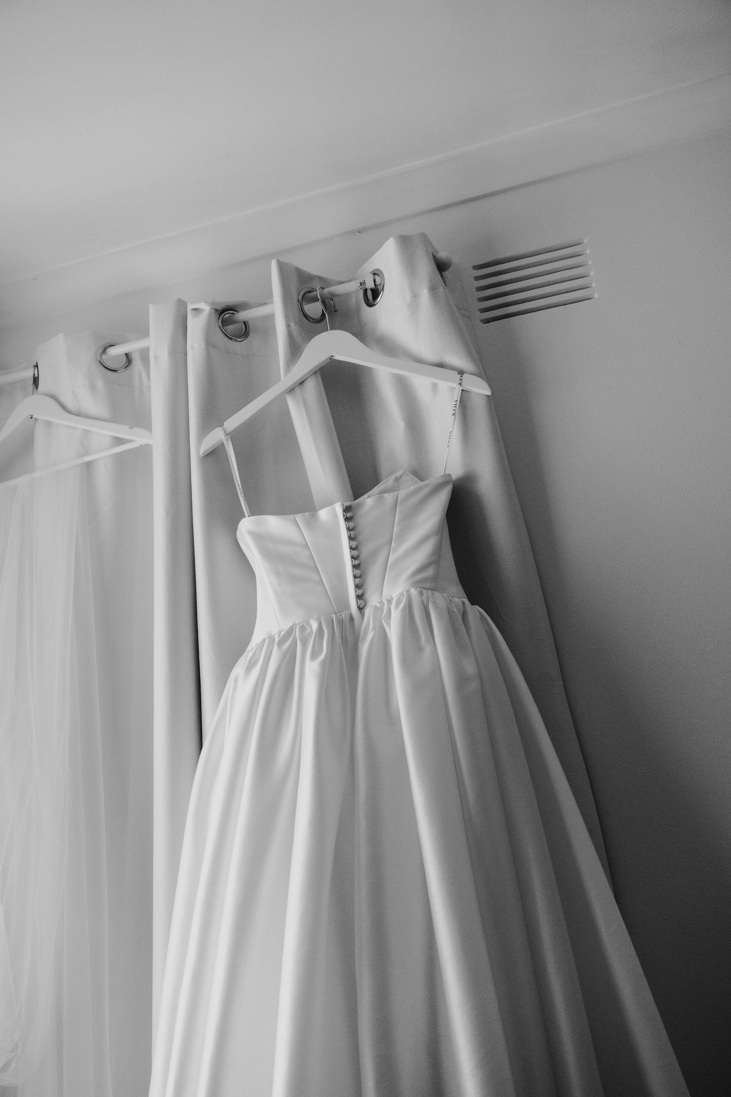 Black and white photo of an elegant strapless wedding dress hanging on a curtain rod with satin finish.
