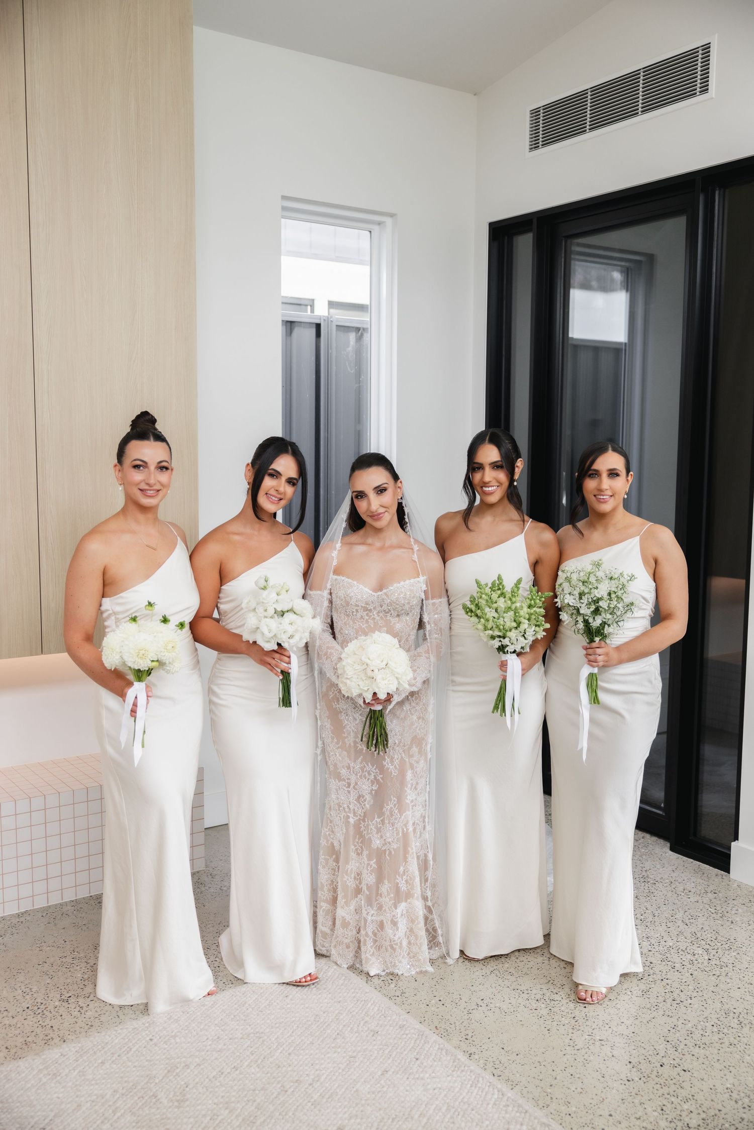 Bride in lace gown with four bridesmaids in white satin dresses holding bouquets of white flowers indoors.