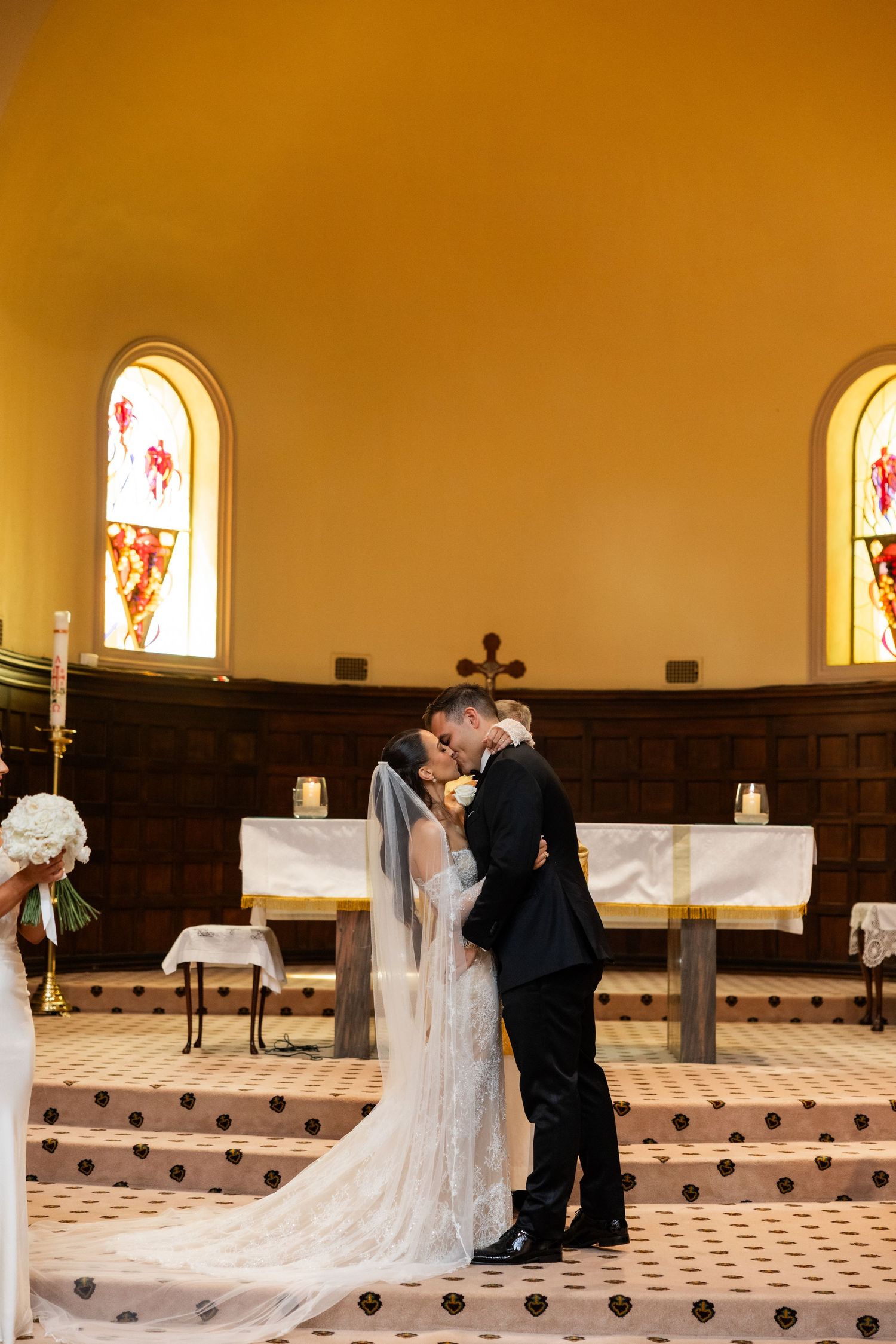 Bride and groom sharing first kiss at church altar with stained glass windows and candlelight.