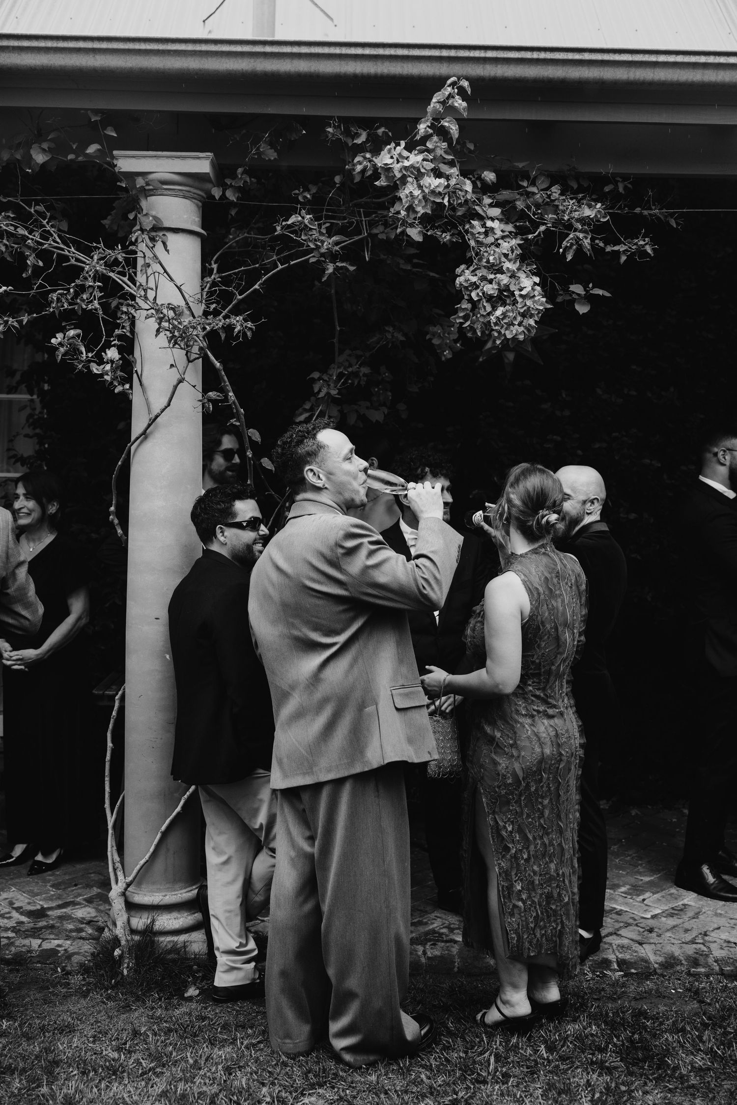 Black and white photo of wedding guests socializing outdoors near a column, with one man drinking from a bottle.