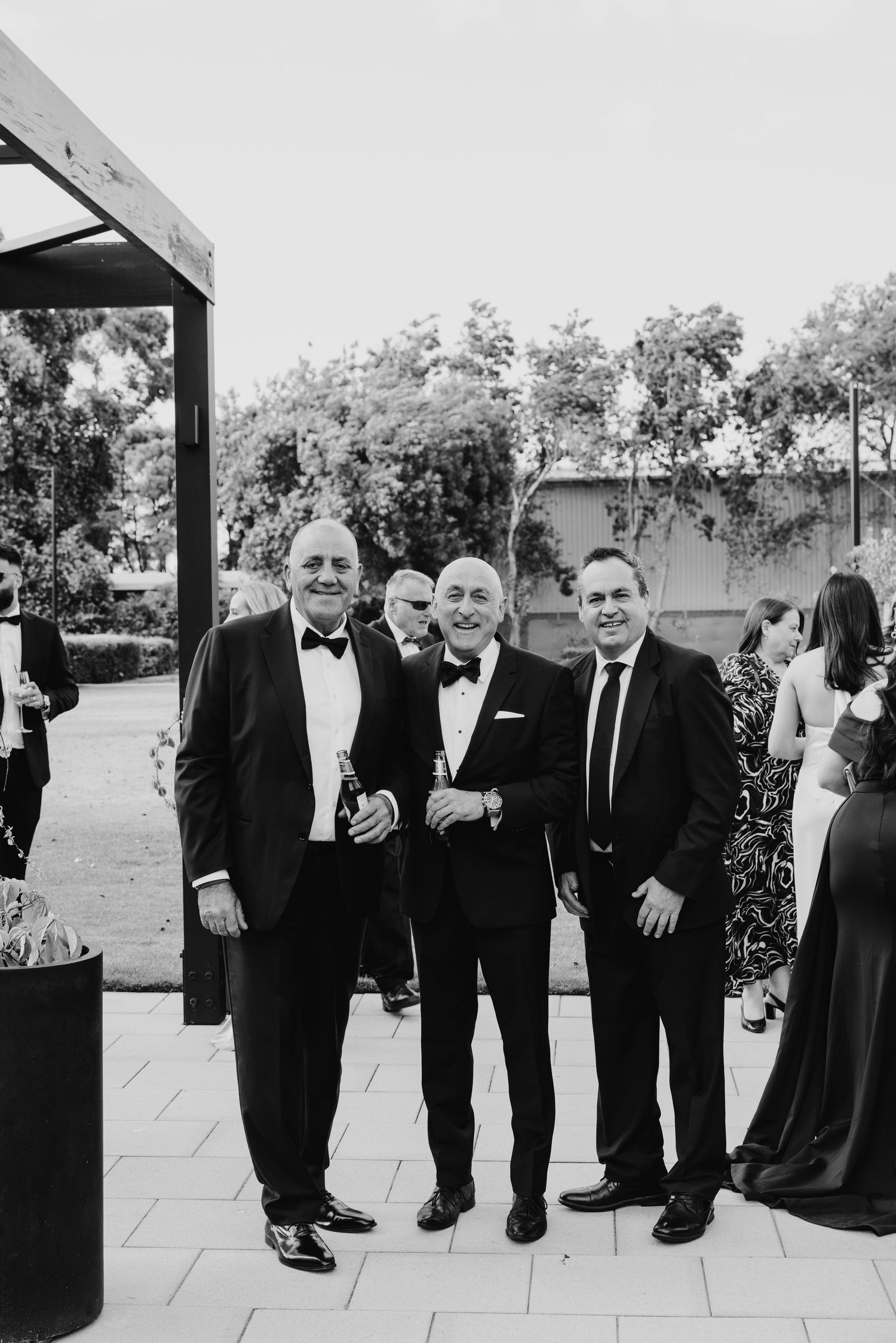 Three men in formal black tie attire pose together smiling at an elegant outdoor event.