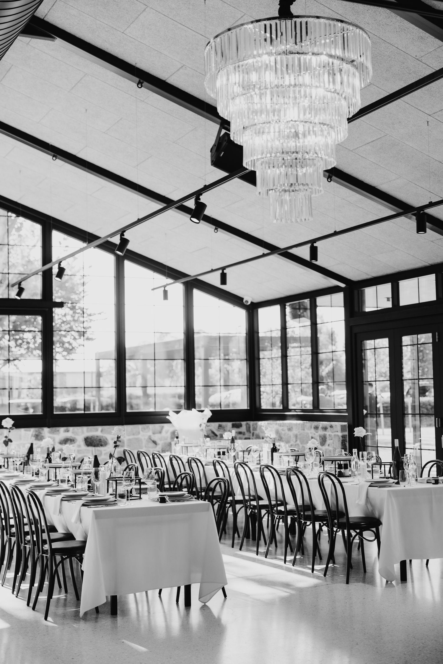 Elegant black and white photo of a sunlit reception hall with white-draped tables, bentwood chairs, and a crystal chandelier.