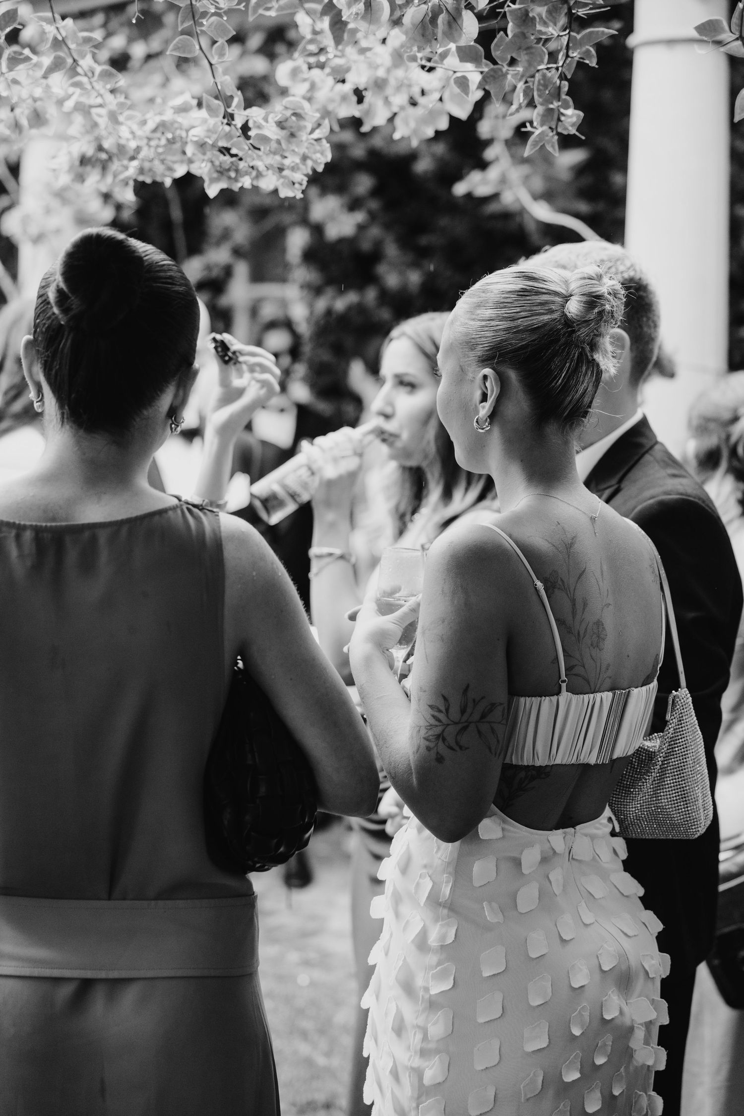 Black and white photo of elegant women in formal dresses socializing outdoors at a wedding reception.