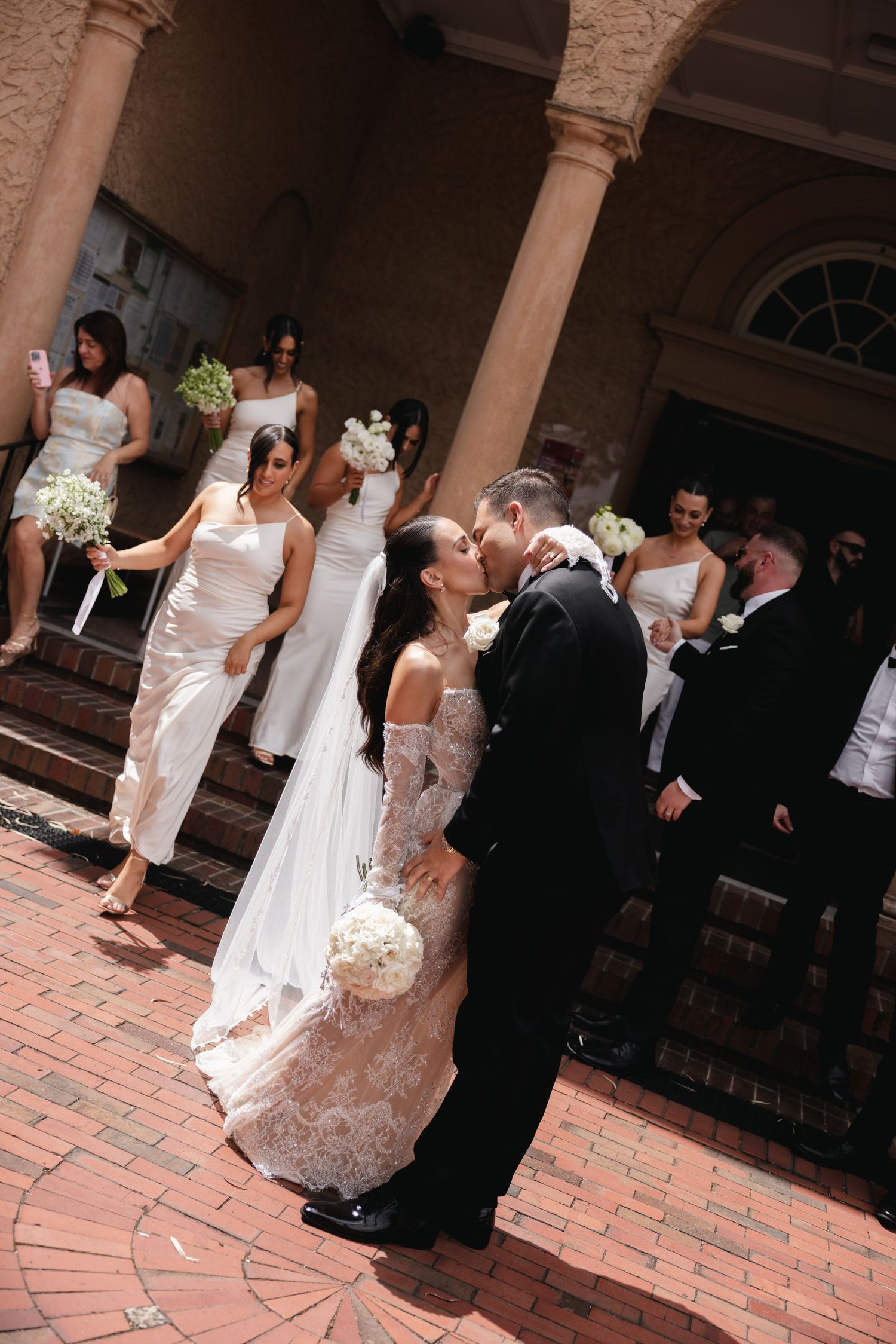 Bride and groom sharing first kiss outside church with wedding party.