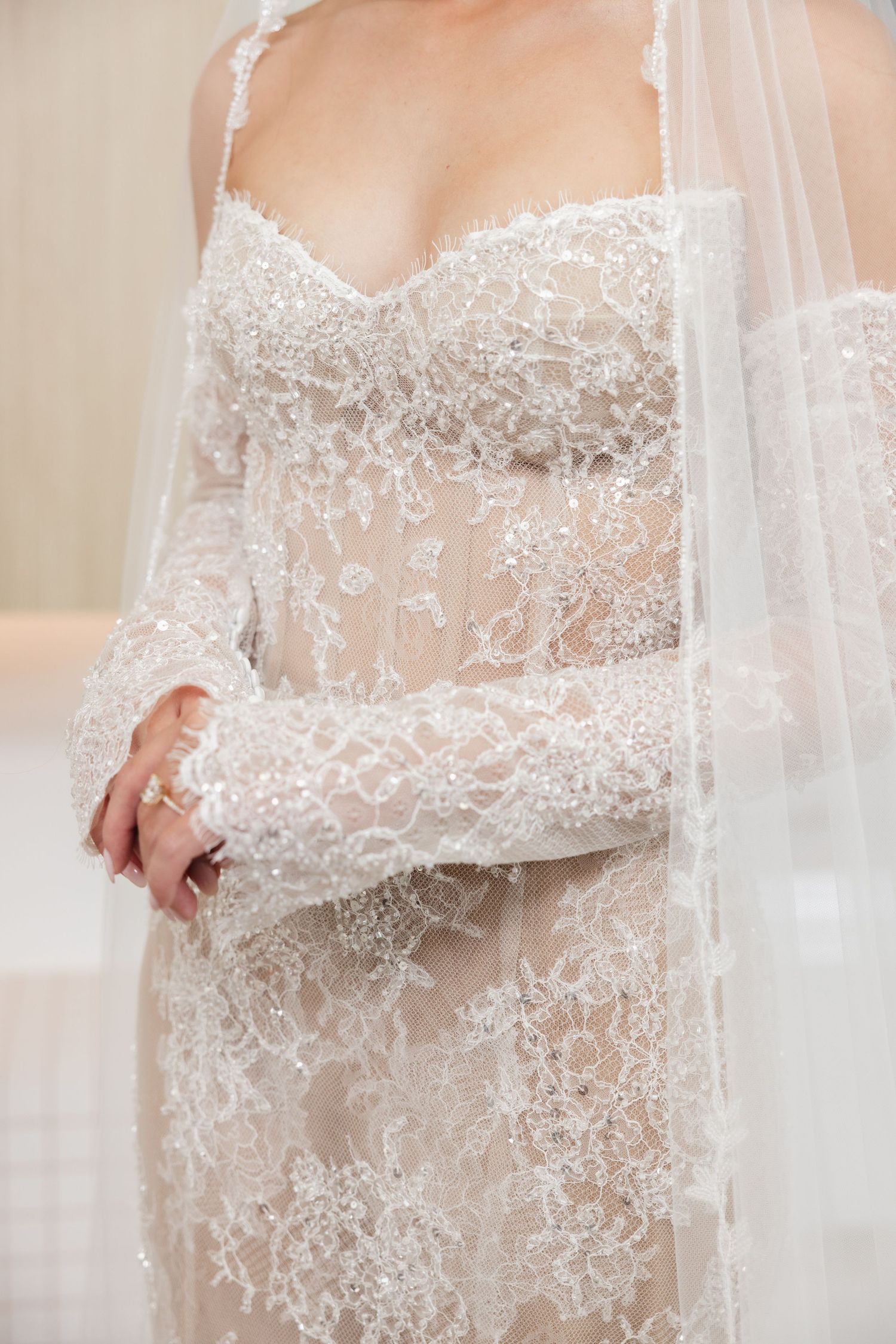 Close-up of bride wearing intricate beaded lace wedding gown with sheer sleeves and flowing veil.