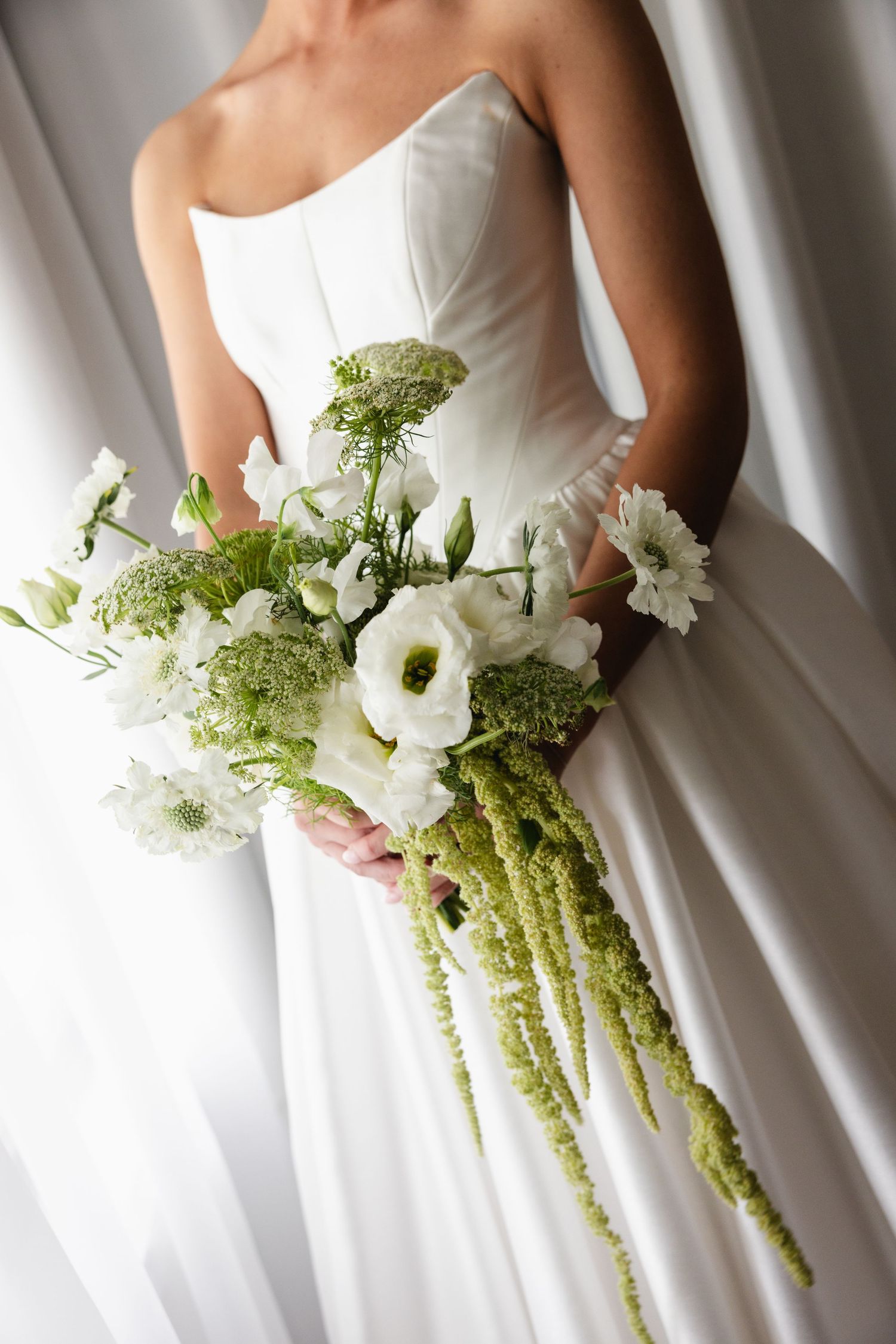 Bride in white strapless gown holding elegant bouquet of white flowers and green amaranthus cascading stems.