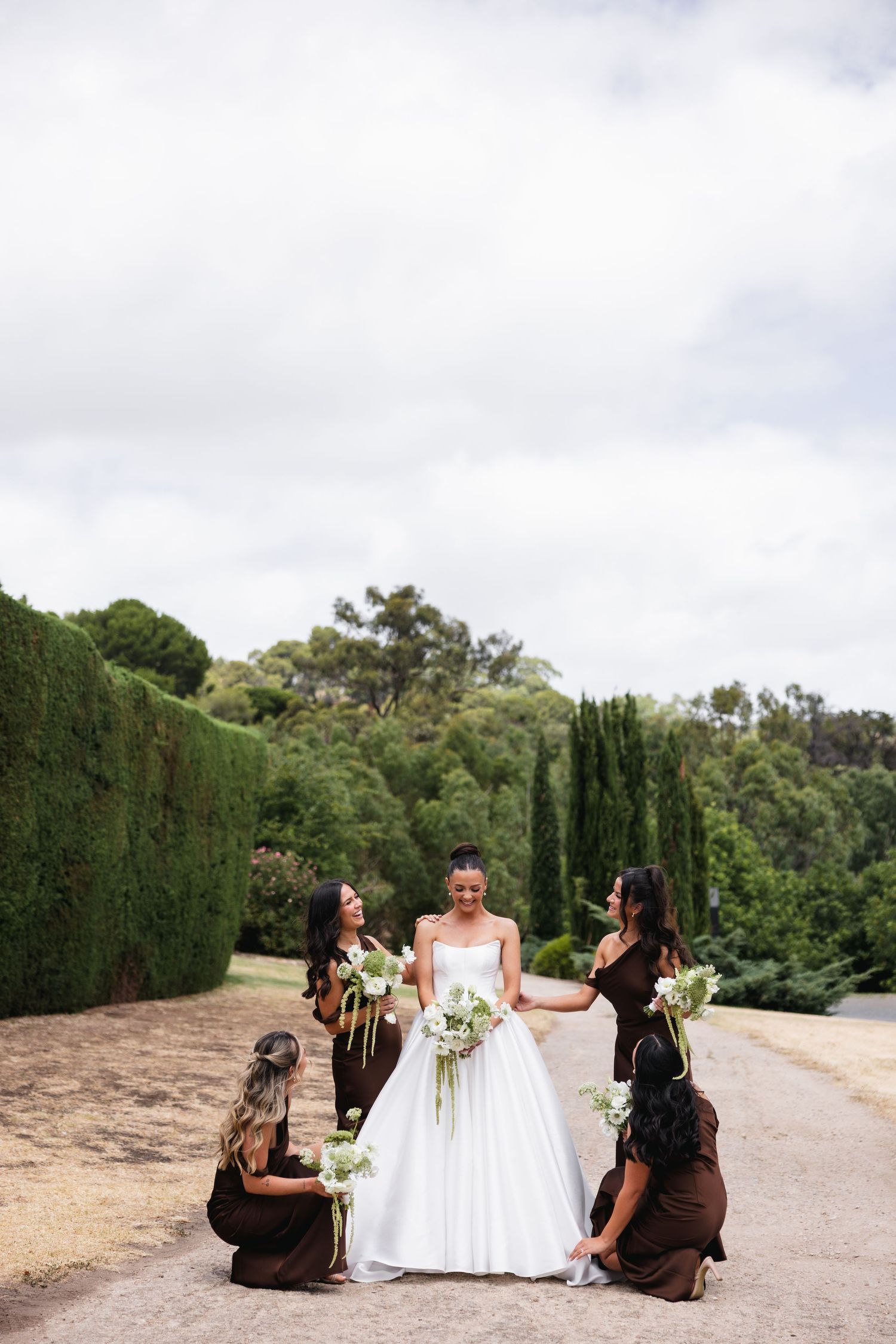 Bride in white gown surrounded by bridesmaids in brown dresses holding white bouquets on a garden path.