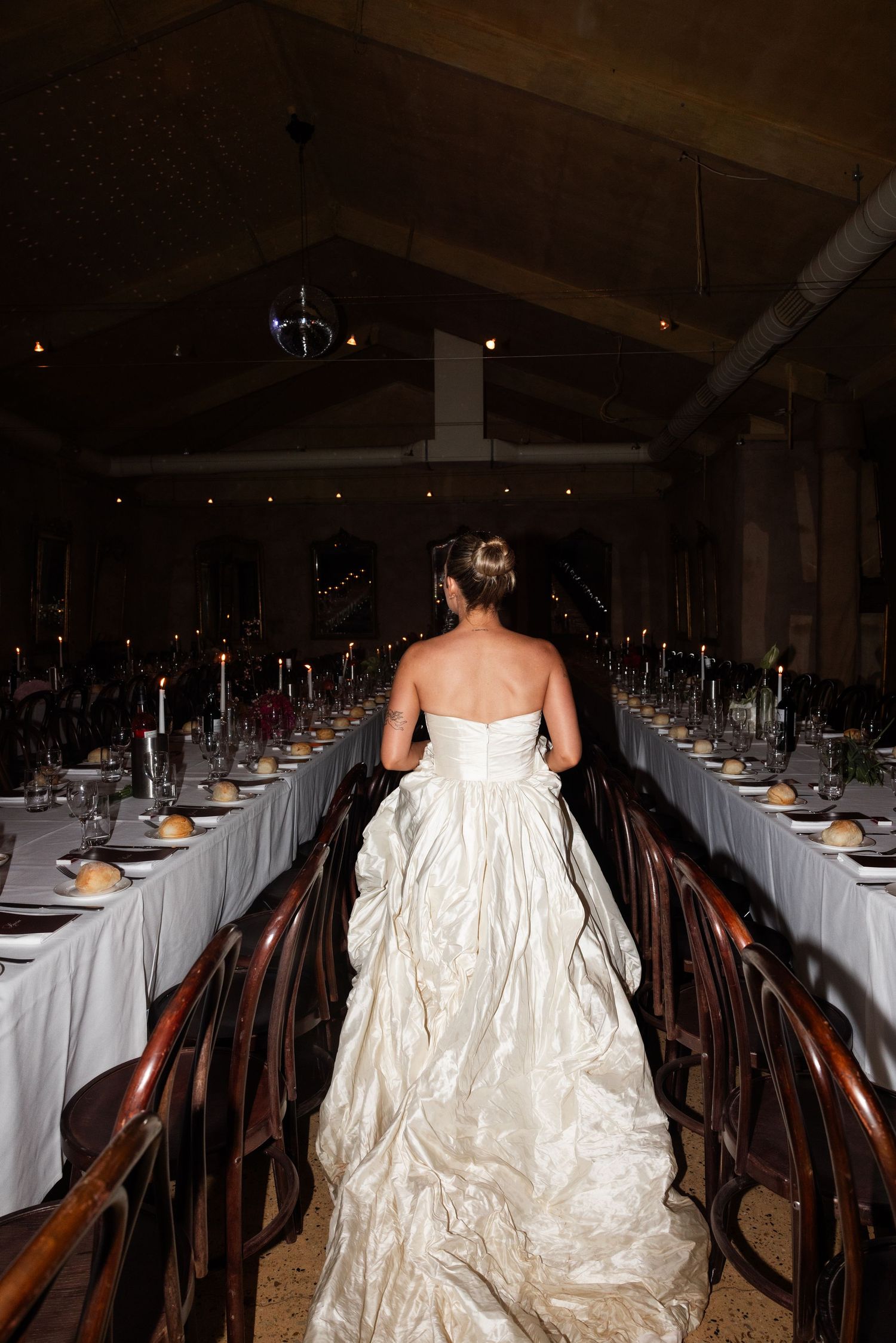 Bride in elegant white ball gown walks between long reception tables set with candles in a dimly lit venue.