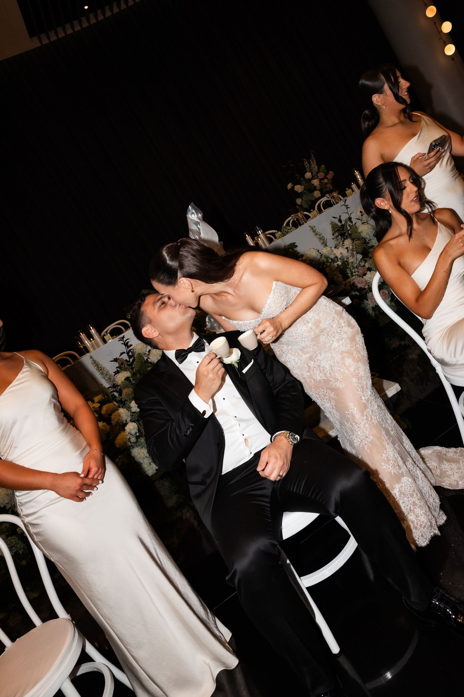 Bride in lace gown kisses groom in tuxedo during wedding reception while bridesmaids look on nearby.