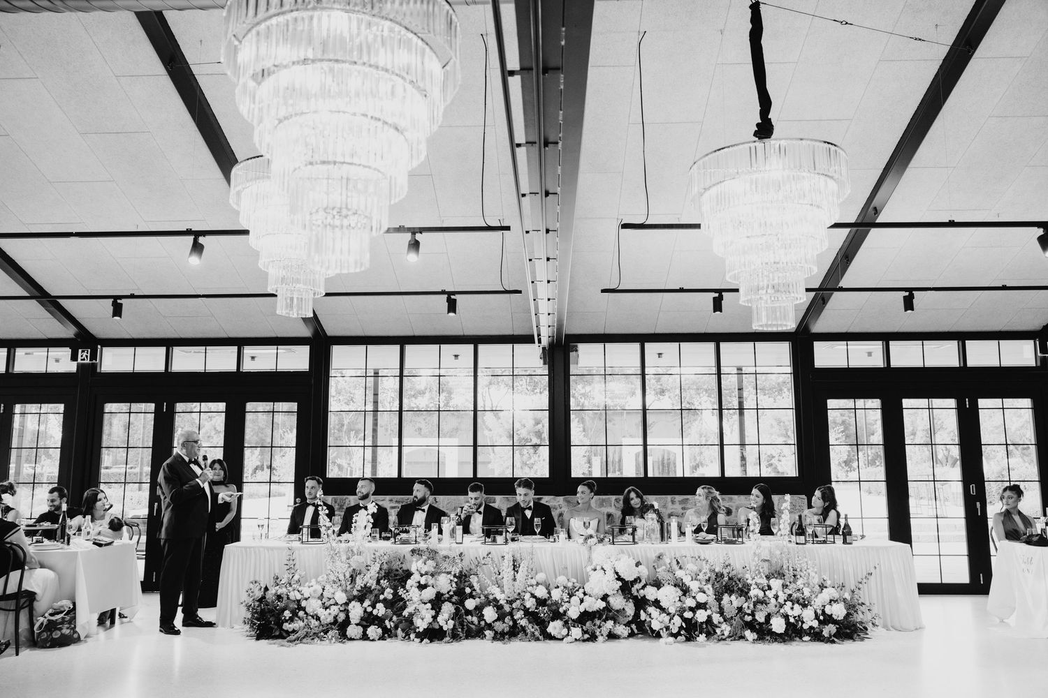 Black and white wedding reception in elegant venue with chandeliers, floral head table, and guests seated beneath large windows.