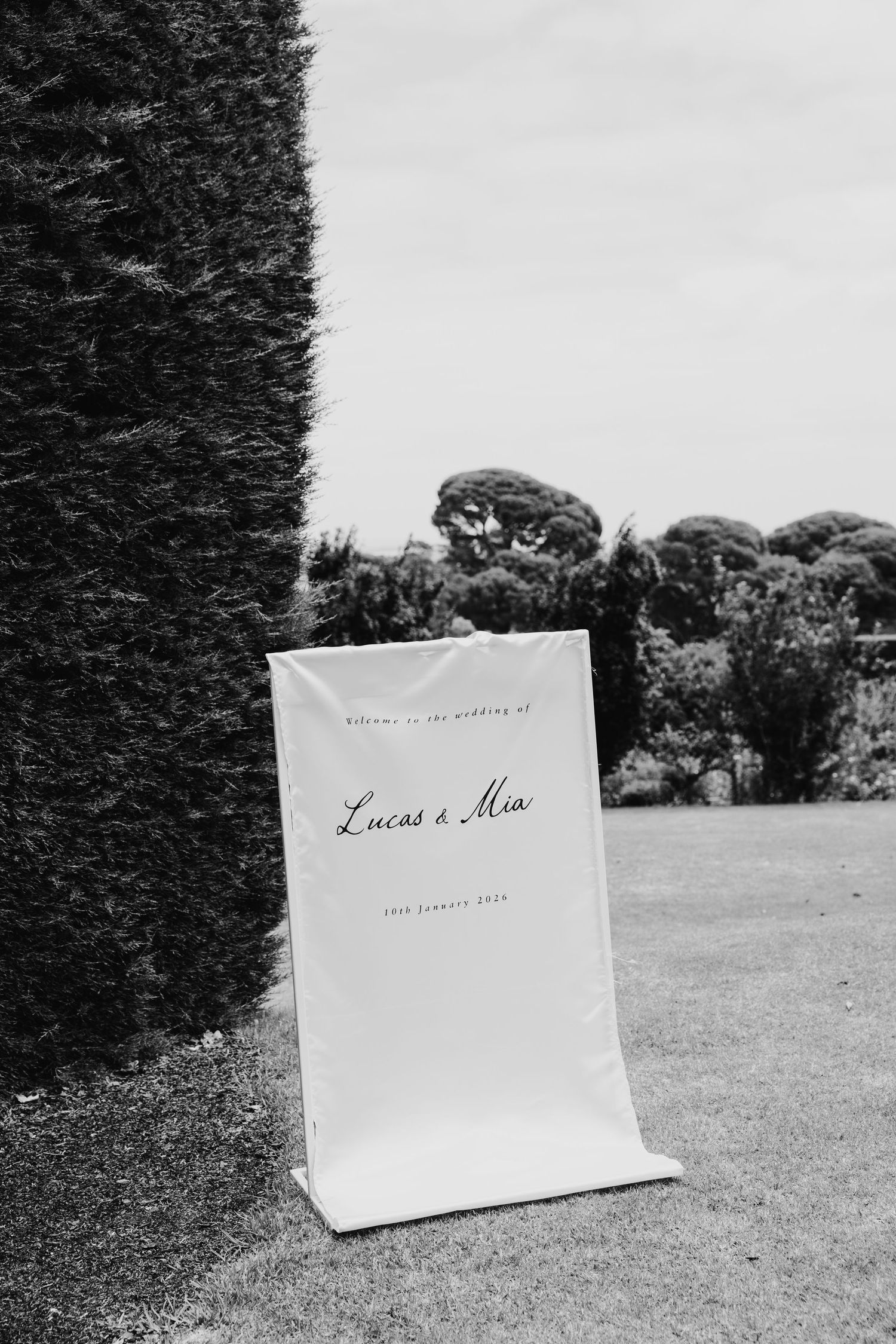 Black and white photo of a wedding welcome sign for Lucas and Mia, leaning against a tall hedge outdoors.