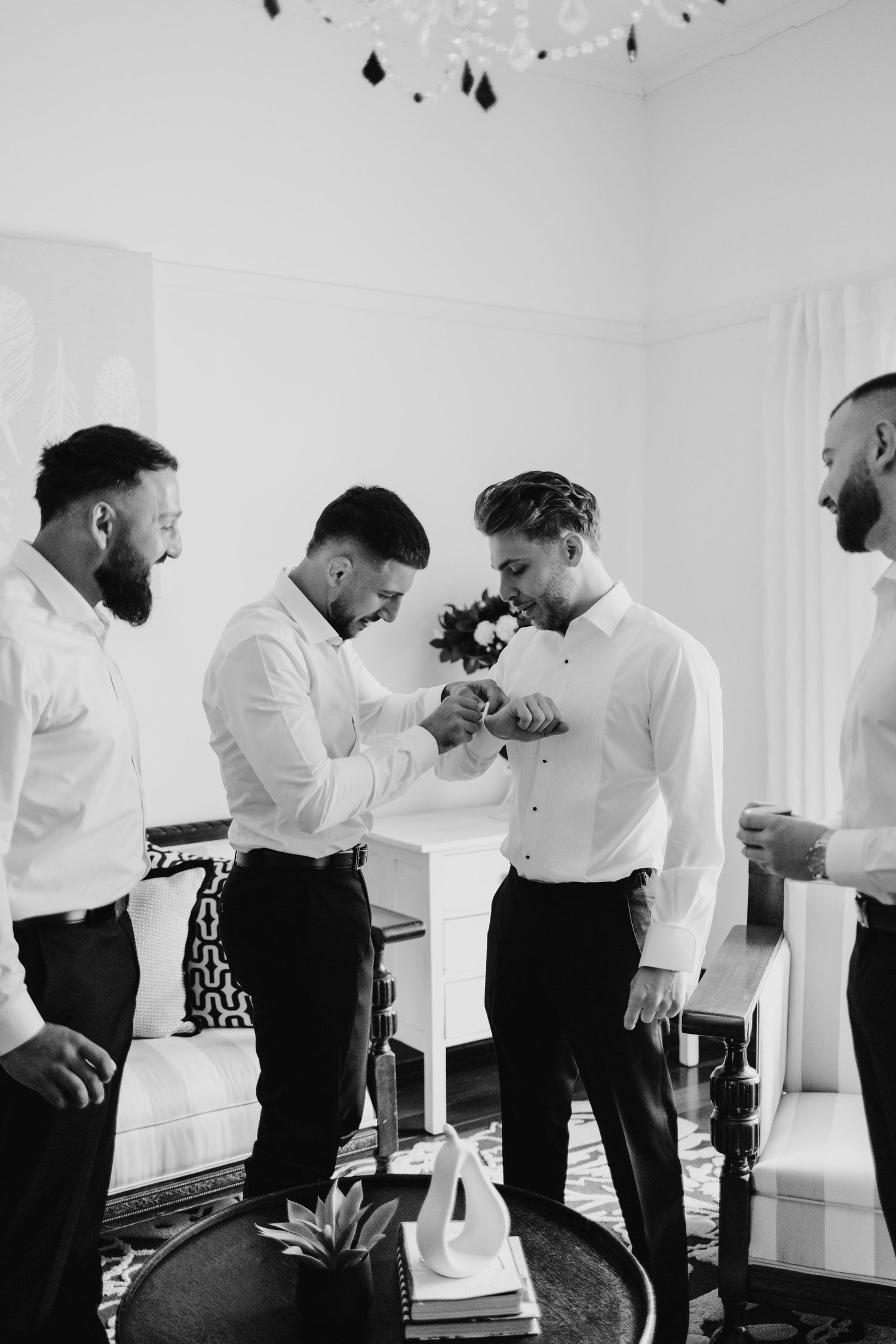 Groomsmen helping groom with cufflinks in bright white room.
