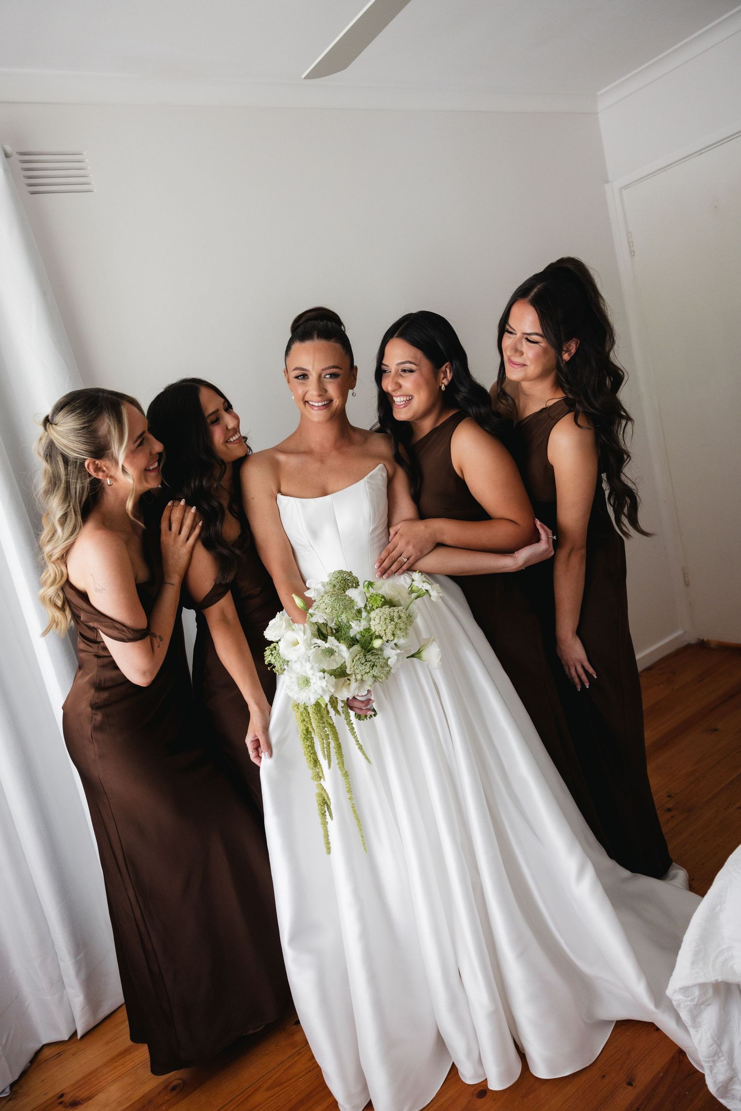 Bride in white gown holds green bouquet surrounded by four bridesmaids in brown dresses indoors.