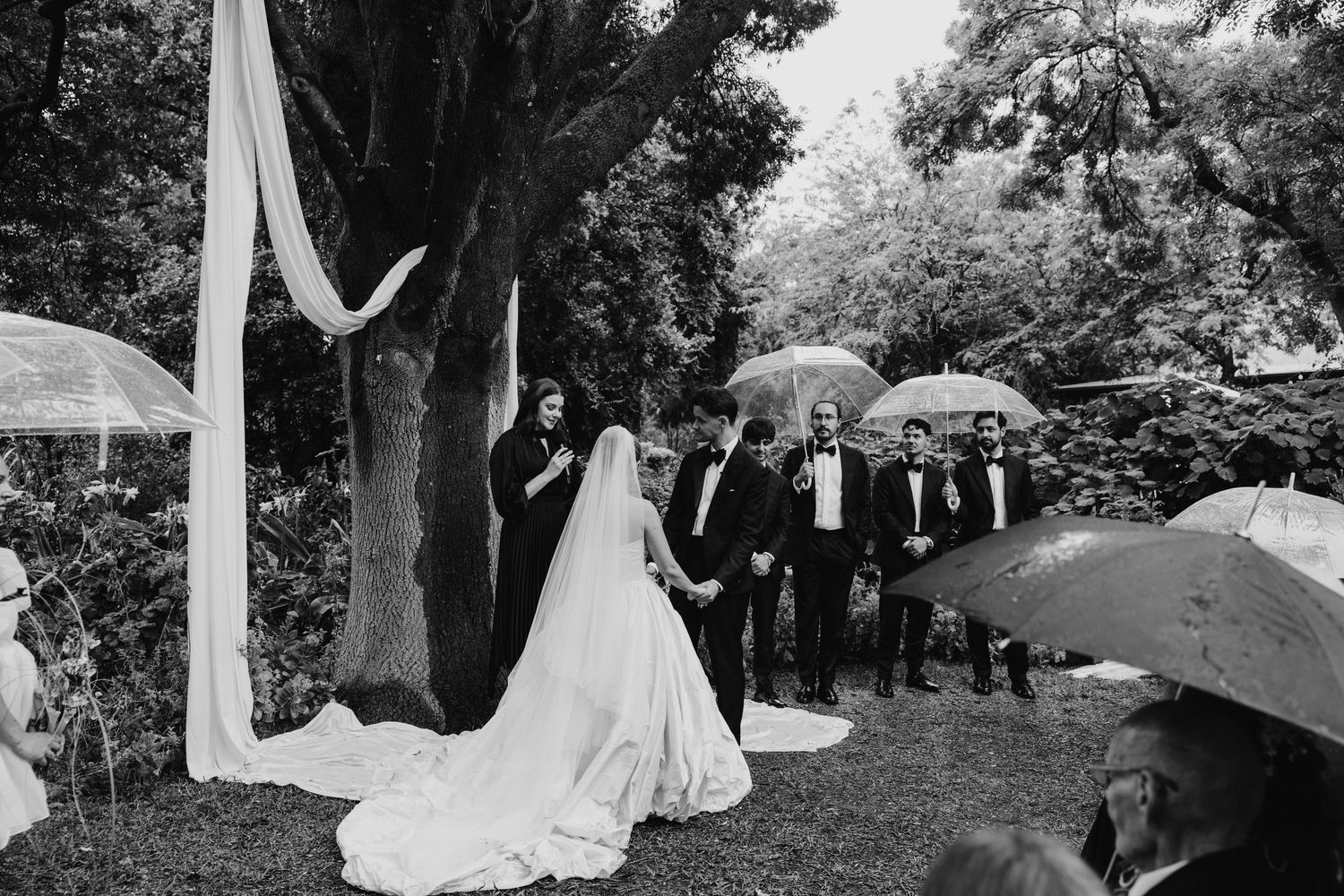 Black and white outdoor wedding ceremony in a garden, bride in gown, guests holding umbrellas in the rain.