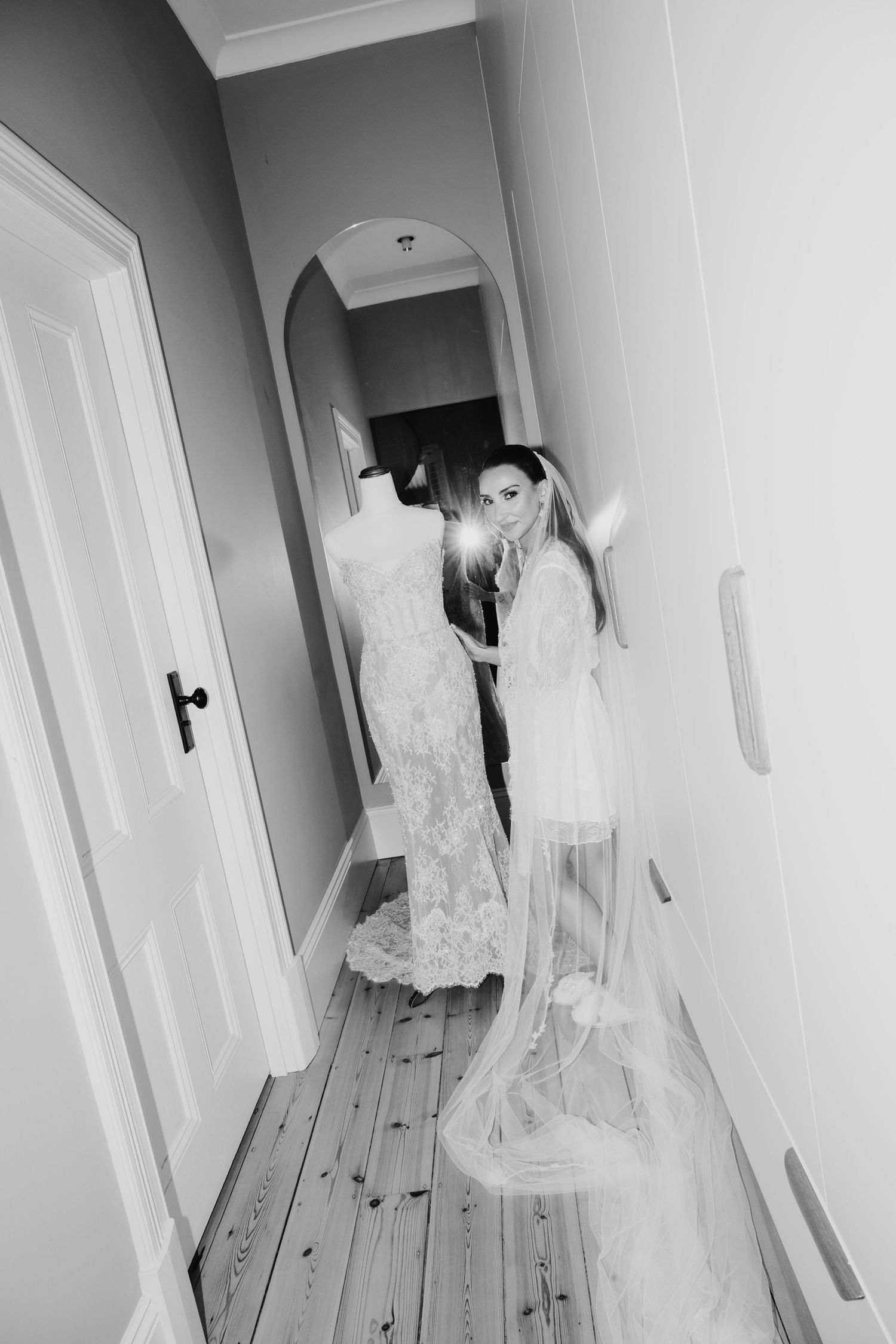 Black and white photo of a bride in a lace gown and veil standing in a hallway next to her wedding dress on a mannequin.