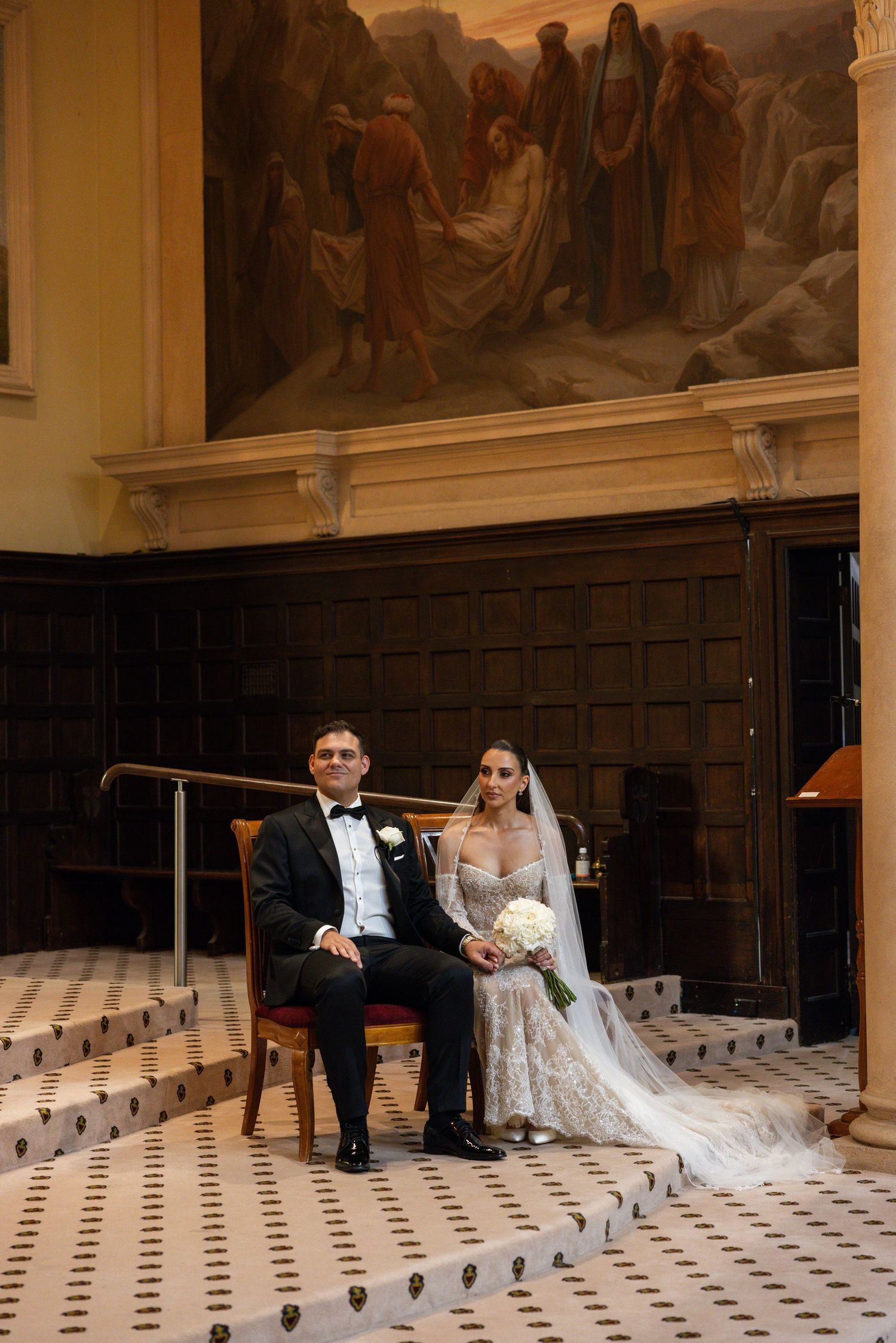 Bride and groom seated at church altar beneath a grand classical painting during wedding ceremony.