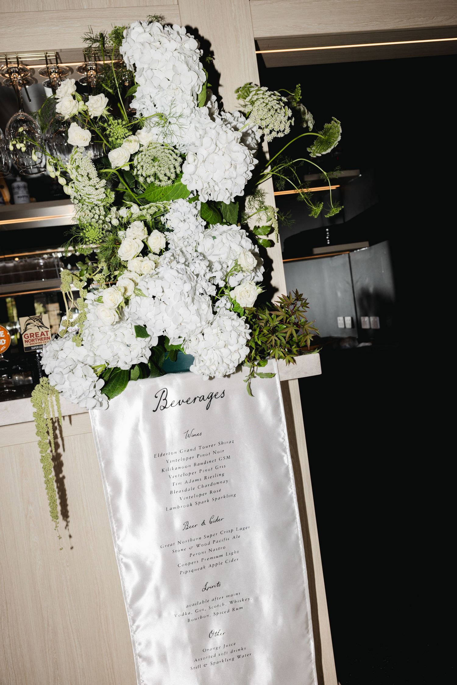 Elegant wedding beverages sign adorned with white hydrangeas and greenery floral arrangement.