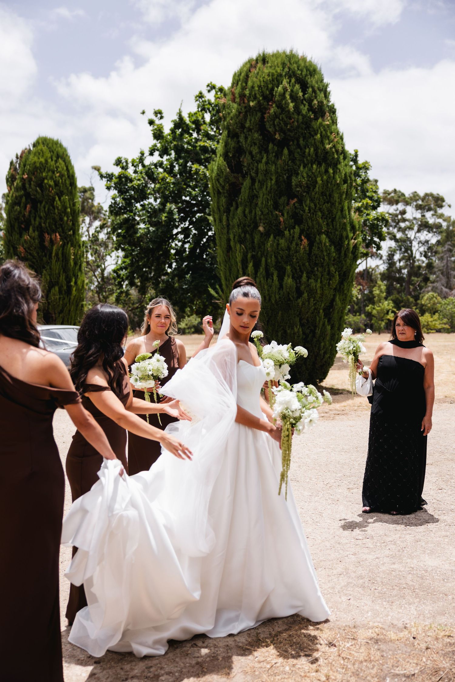 Bride in white ball gown holding bouquet assisted by bridesmaids in black dresses outdoors near cypress trees.