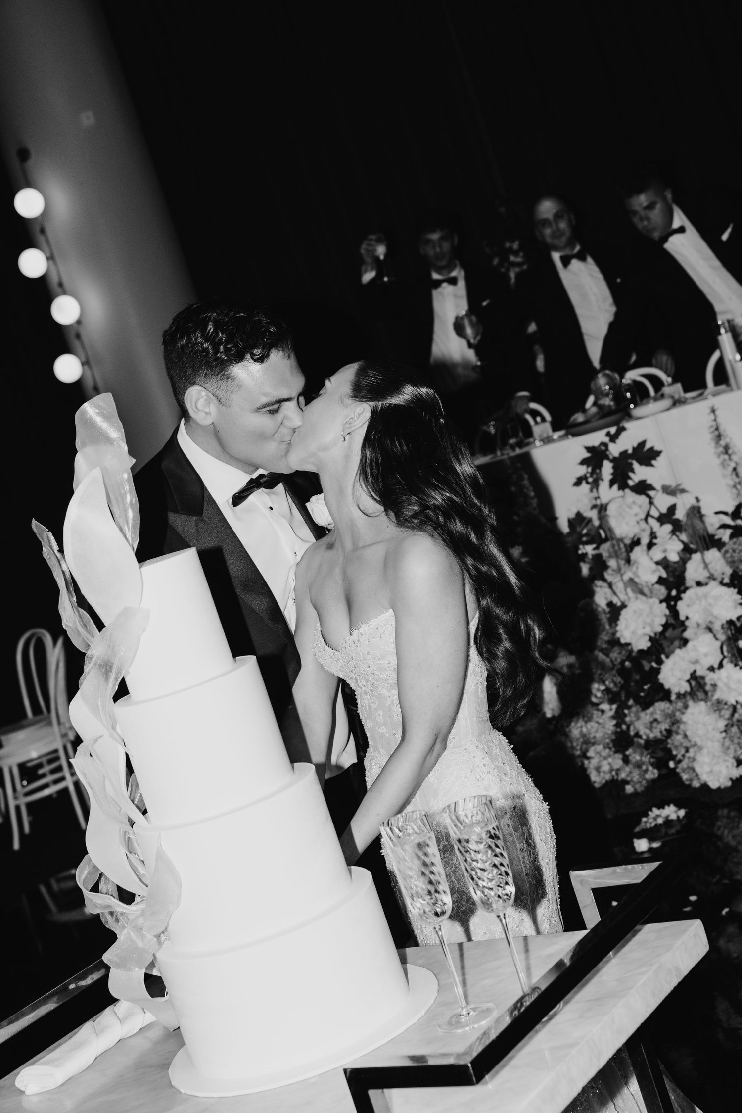 Bride and groom share a kiss beside a tall white tiered wedding cake in black and white photo.