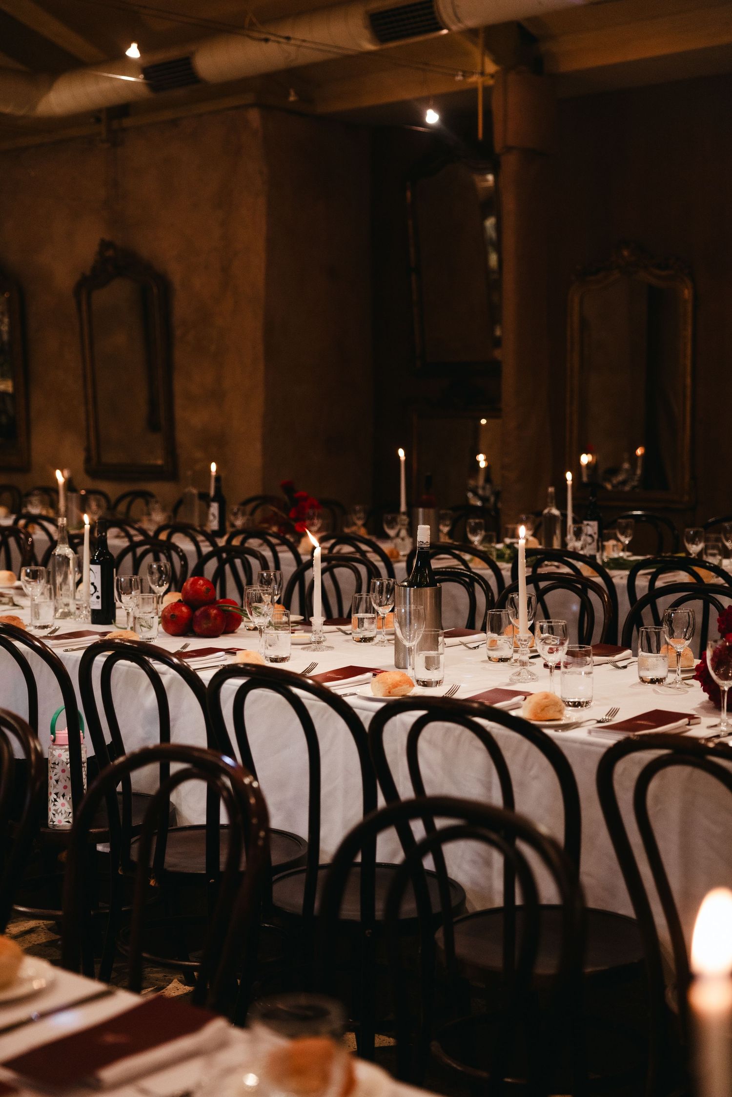 Elegant candlelit wedding reception with long tables, black bentwood chairs, and red floral centerpieces in a warm, dimly lit venue.
