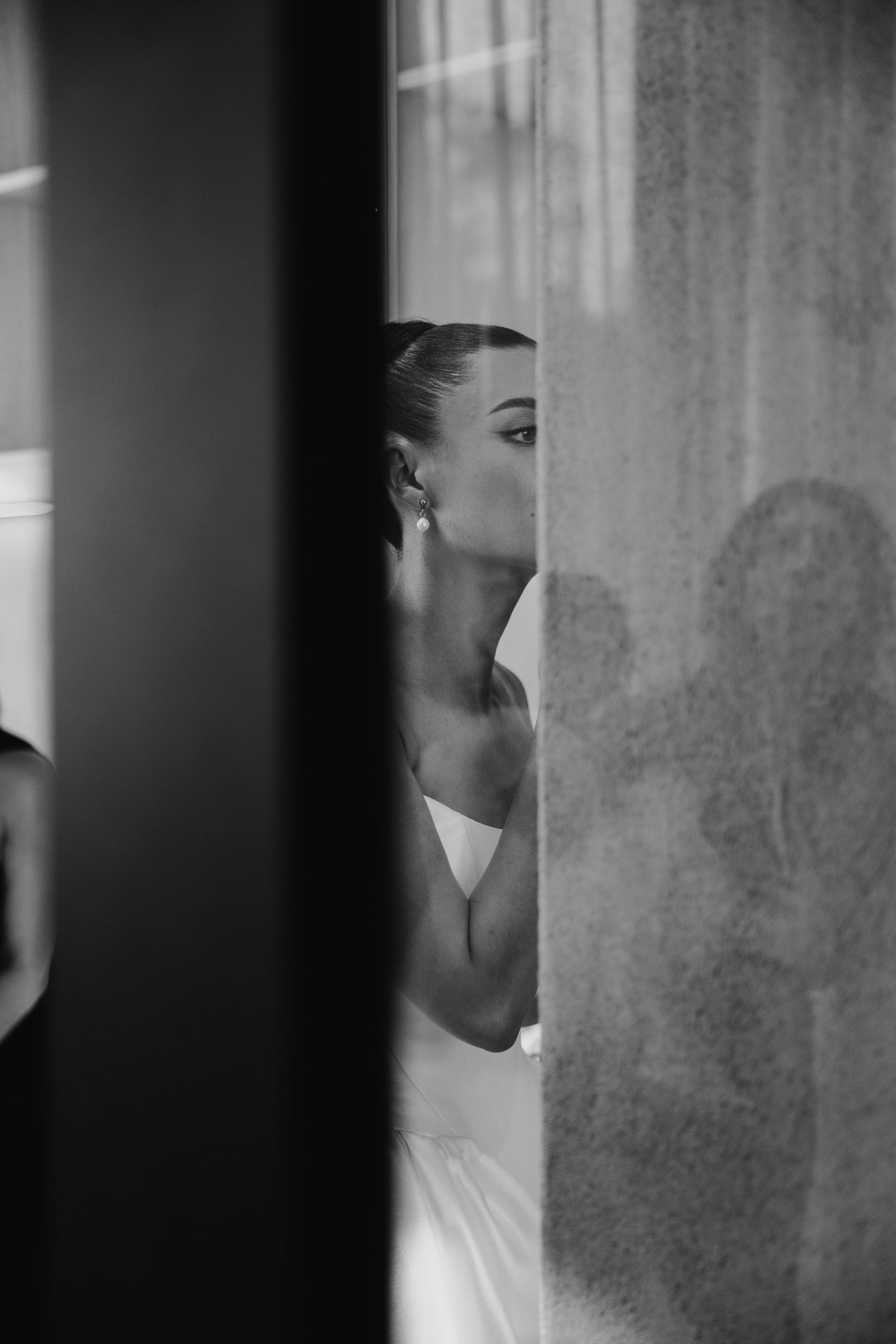 Black and white photo of a bride peeking around a curtain, wearing a strapless gown and elegant earrings.