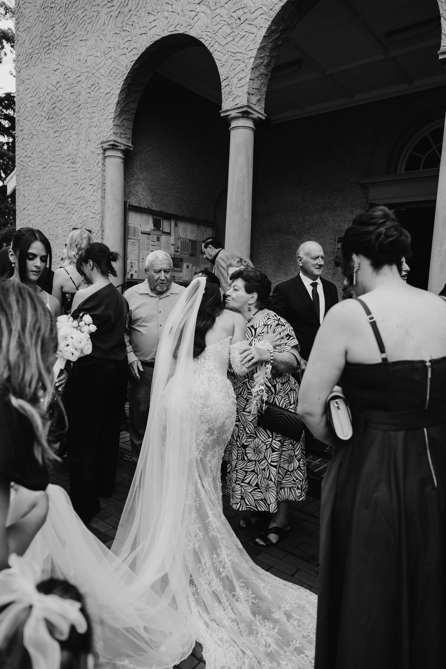 Bride in lace gown with veil greeting elderly guest outside church.