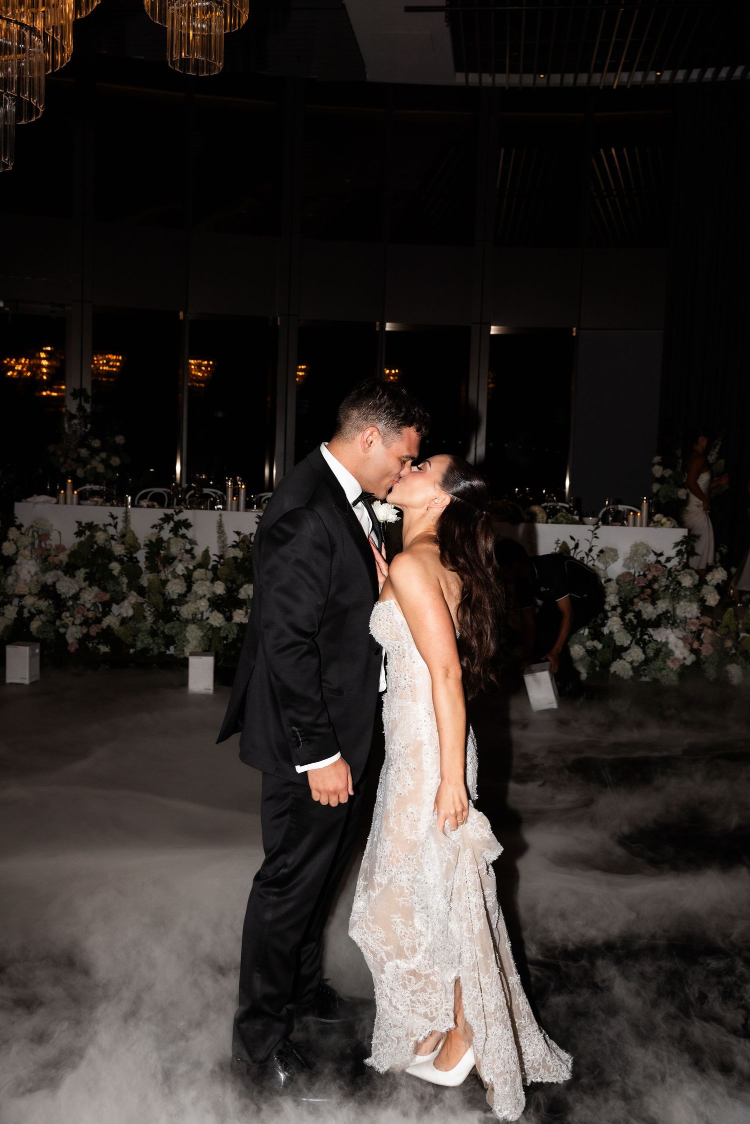 Bride and groom share a romantic first dance kiss surrounded by elegant floral arrangements and dreamy fog at their wedding reception.