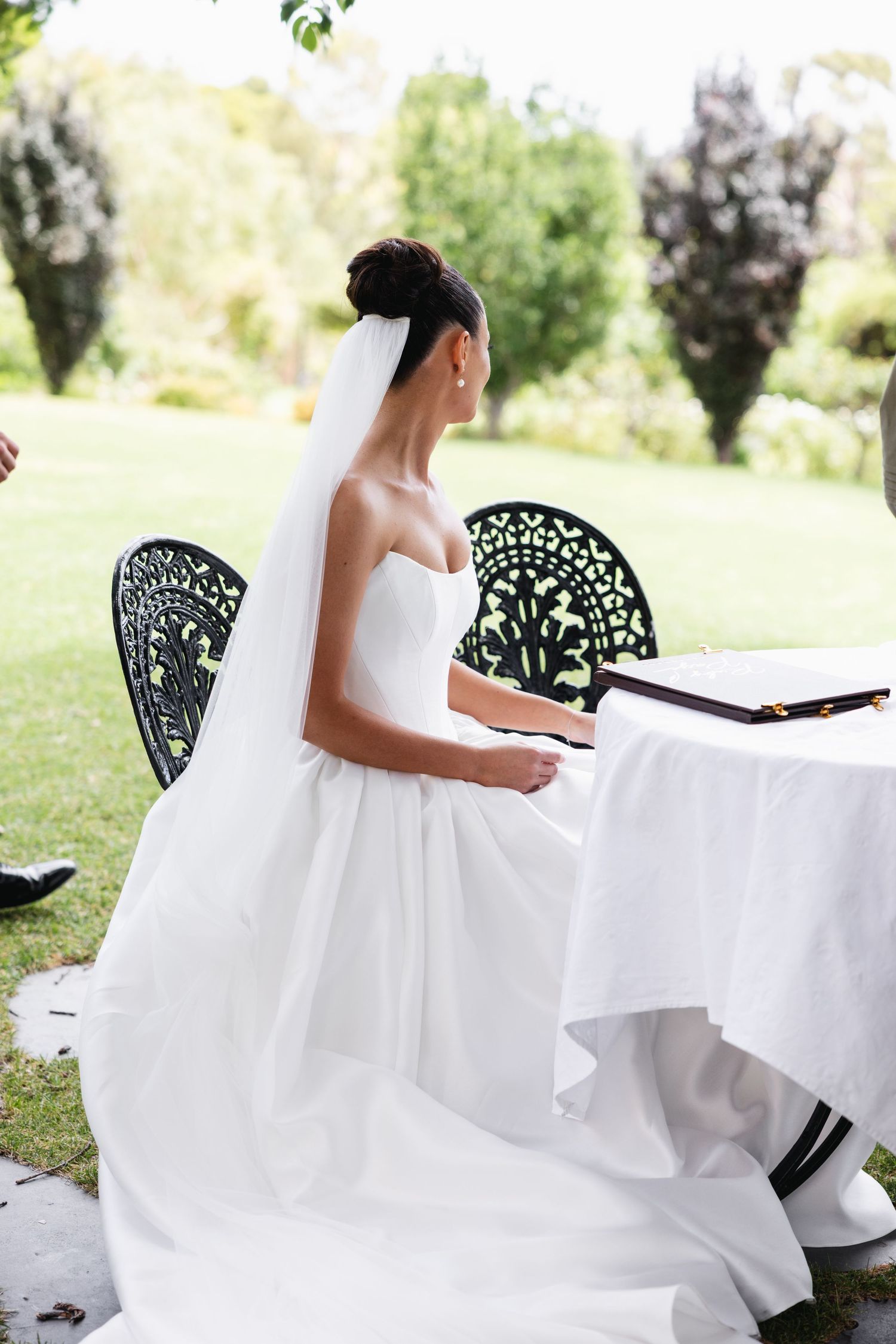 Bride in white strapless gown and veil sits at outdoor table signing marriage register in garden setting.