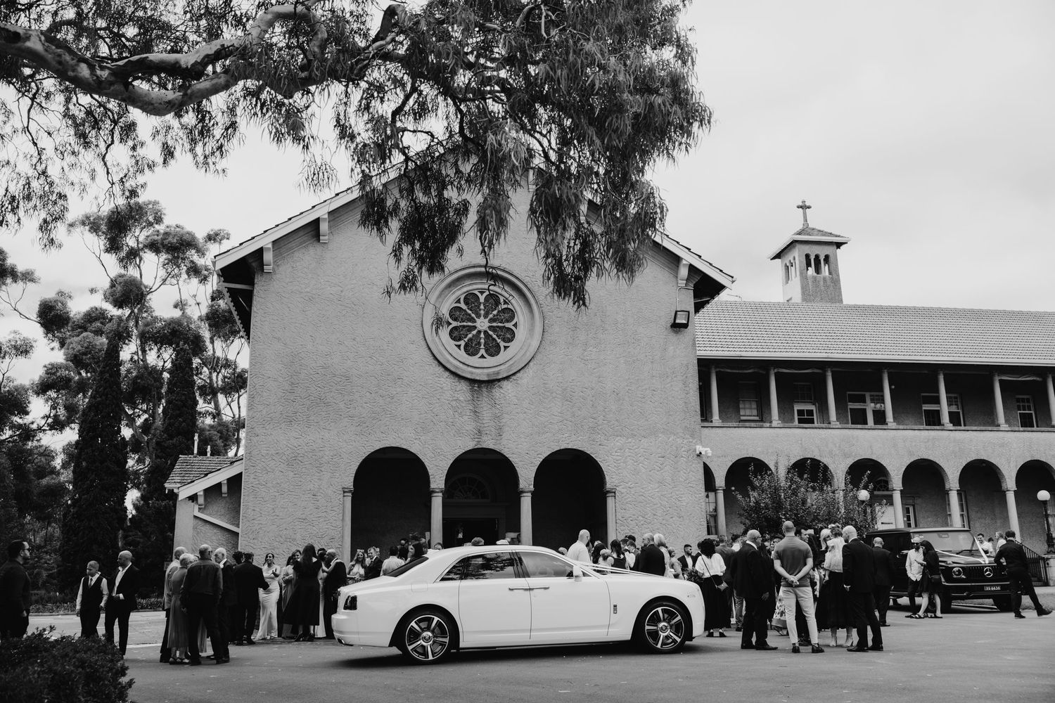 Black and white photo of a white Rolls-Royce outside a church with wedding guests gathered for a ceremony.