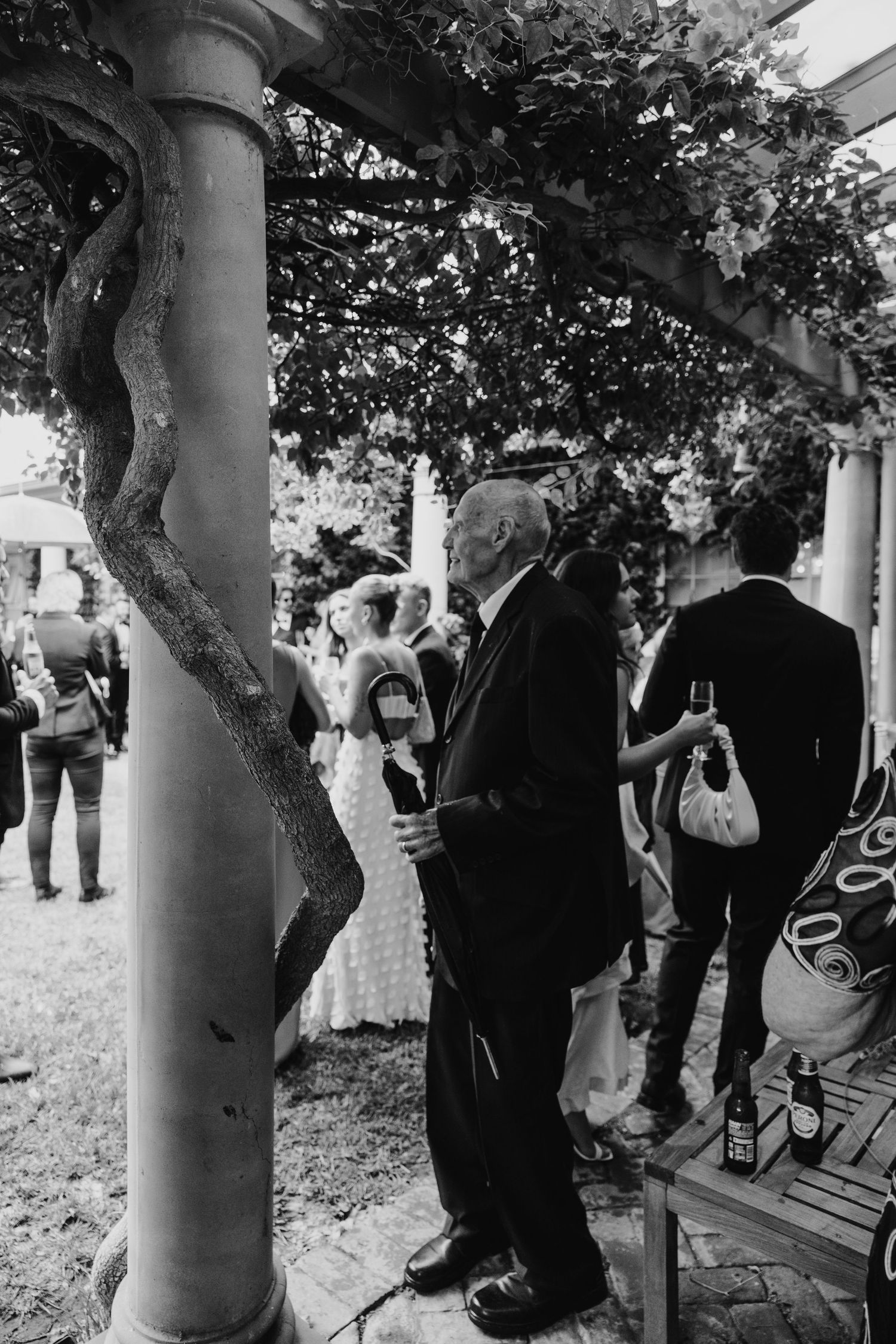 Black and white photo of elderly man in suit holding drink at outdoor garden wedding reception under pergola.
