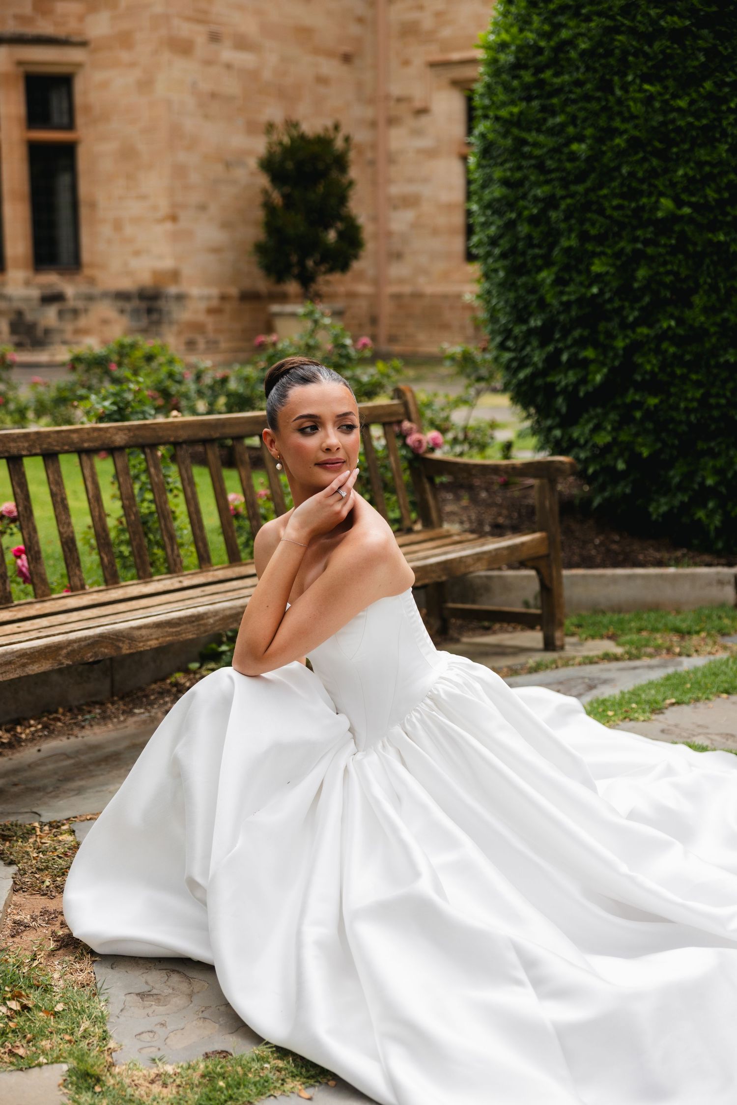 Bride in a strapless white ballgown sits gracefully on stone steps beside a wooden bench in a garden.