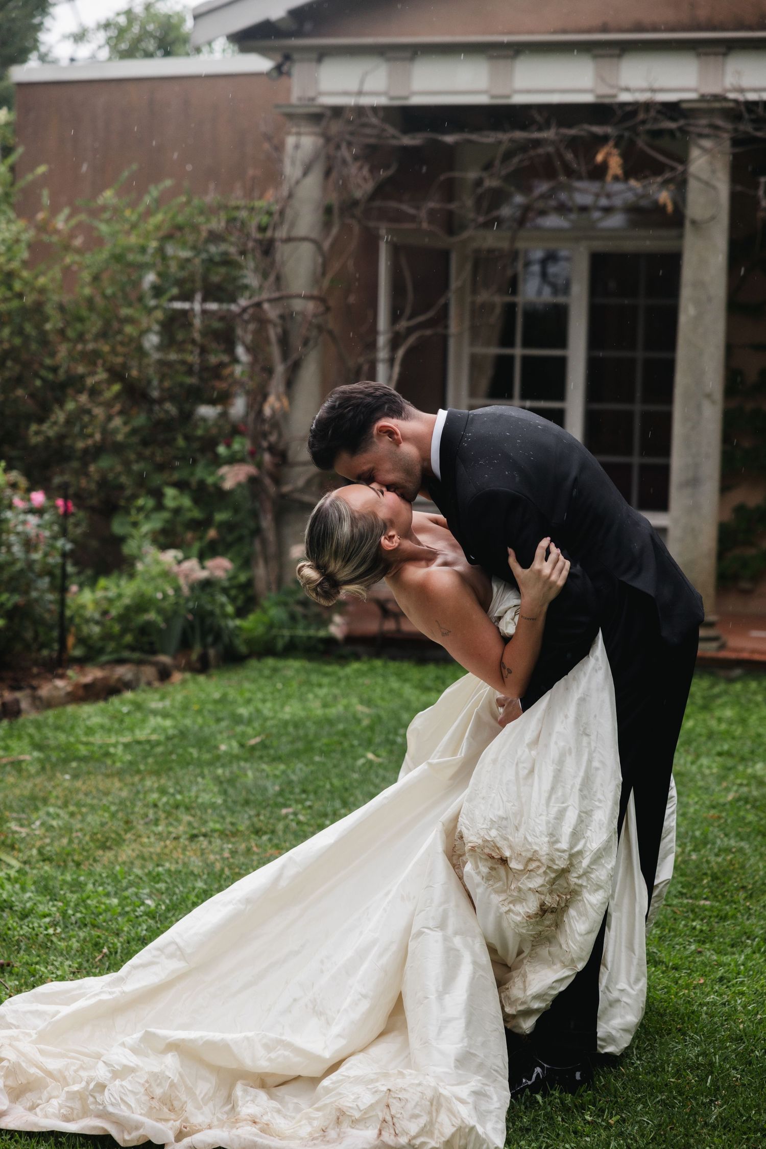 Groom in black tuxedo dips and kisses bride in flowing white gown outdoors on green lawn before elegant estate.