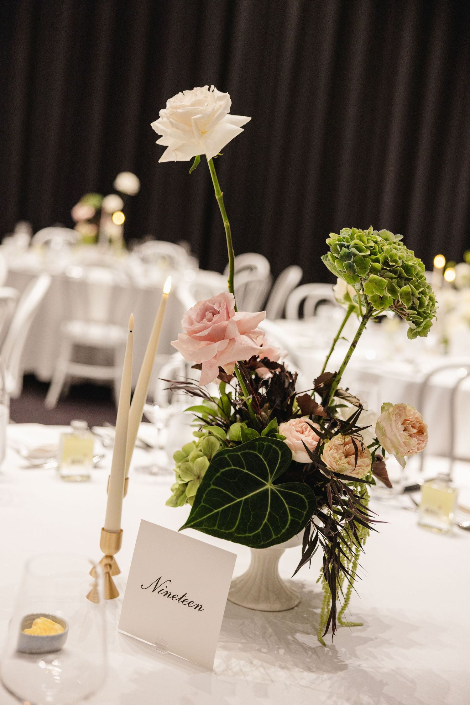 Elegant wedding table centerpiece with white and pink roses, green hydrangeas, and a table number card reading Nineteen.