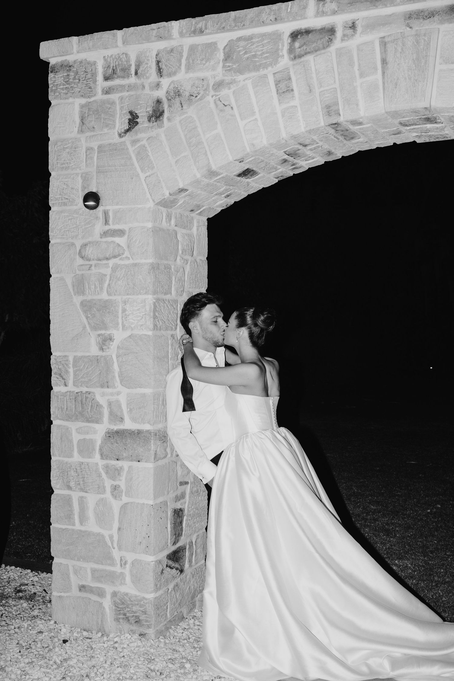 Black and white photo of a bride and groom kissing under a stone arch at night on their wedding day.
