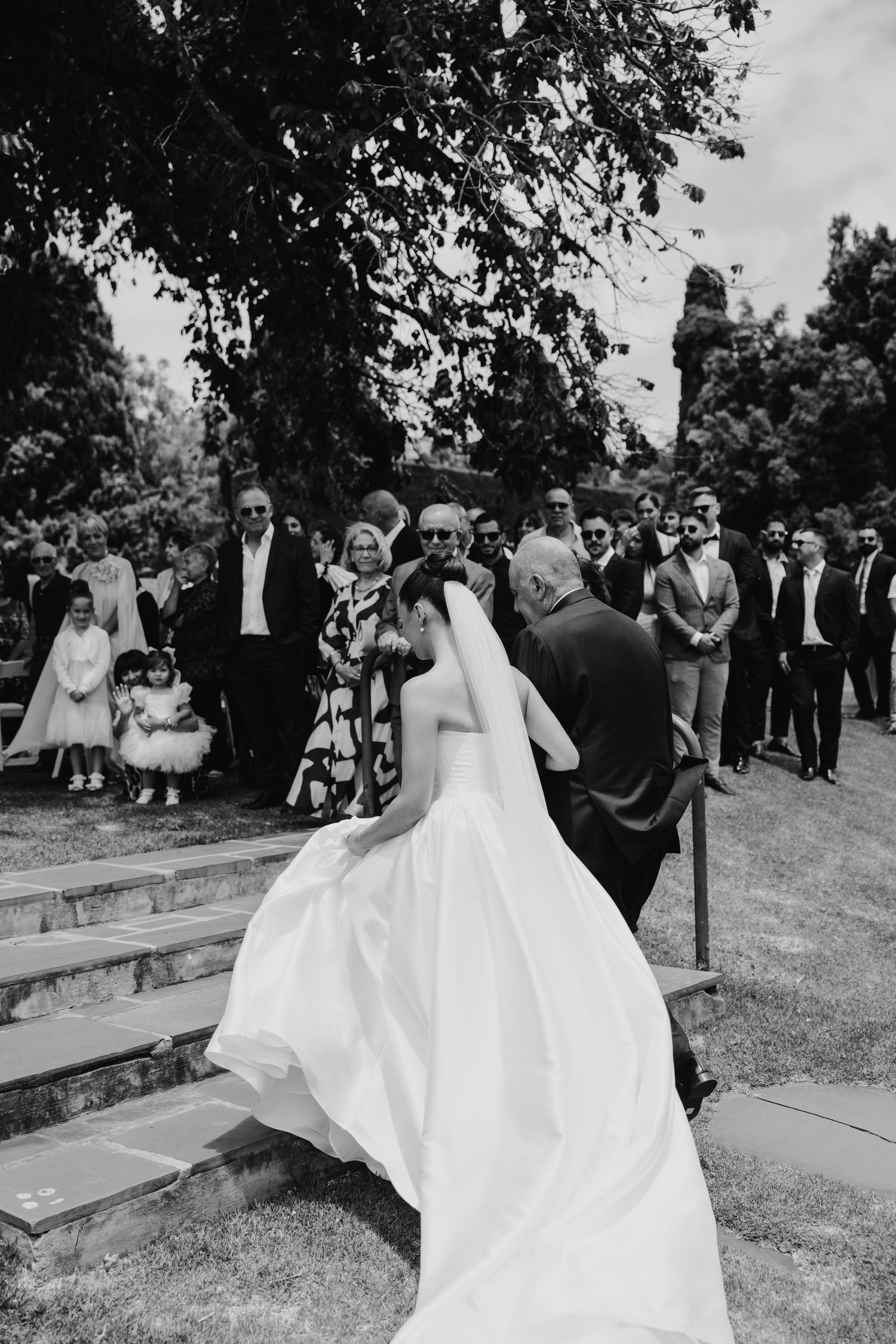 Black and white photo of a bride in a flowing white gown walking up steps with guests gathered outside at a wedding.